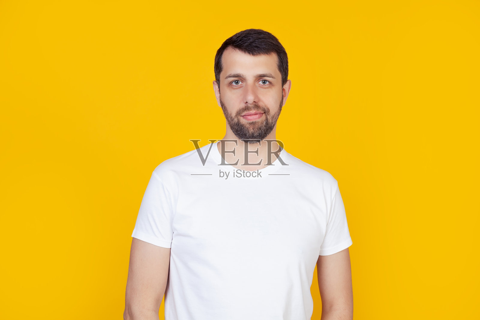 一个留着胡子的年轻人穿着白色的t恤开心的看着镜头微笑。正面的人站在孤立的黄色背景上照片摄影图片