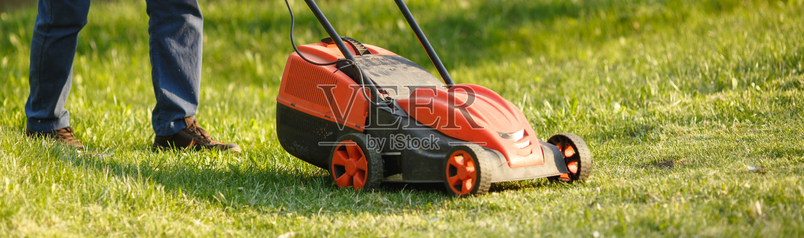 在日落时分，工人在绿色的院子里割草。男人拿着电动割草机，割草。修剪花园的园丁。照片摄影图片