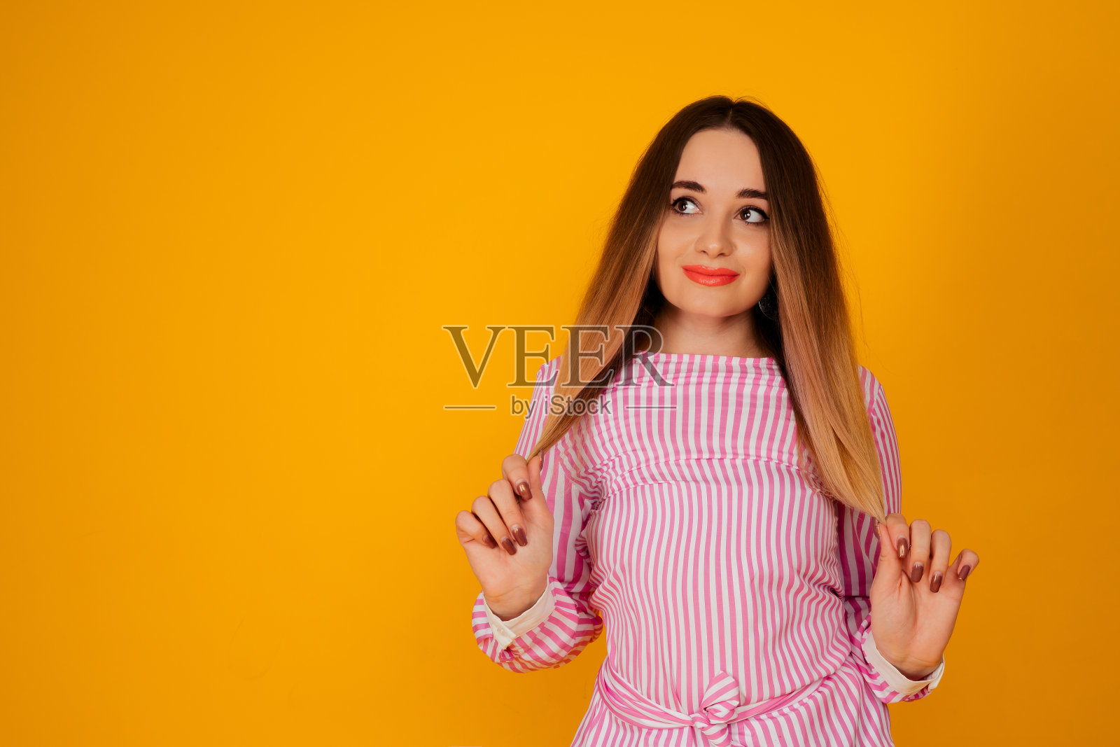 肖像害羞漂亮的年轻女子在时尚的粉红色t恤孤立在黄色的背景。她拿起她的头发。照片摄影图片