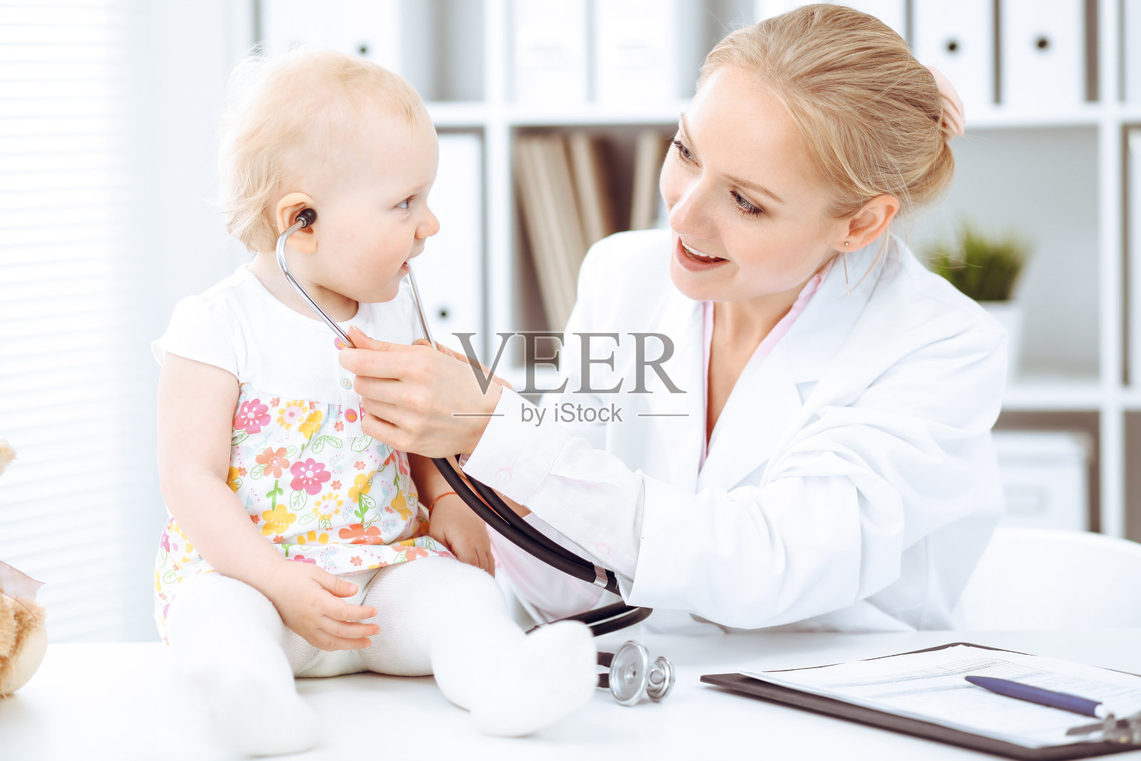 医生和病人在医院里蹒跚学步。小女孩穿着缀有粉红色花朵的连衣裙，正在接受医生的听诊器检查。医学的概念照片摄影图片