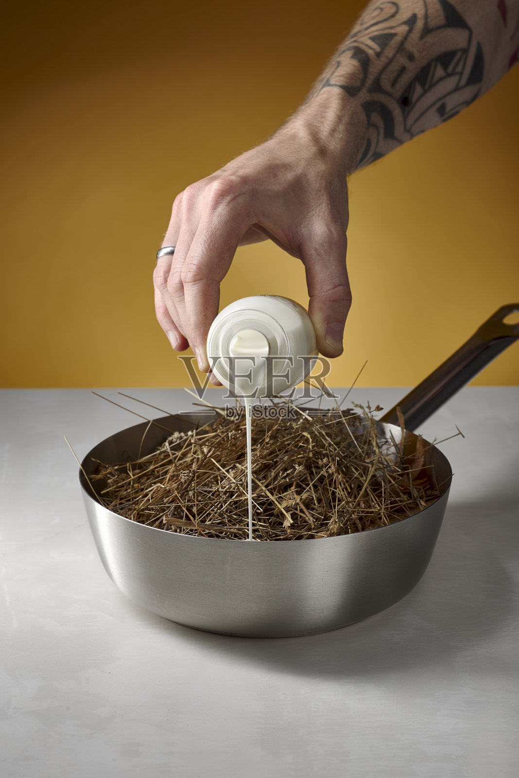 有纹身的手把奶油倒进一个银色的罐子里，罐子里放着烤干草，白色大理石桌子上放着黄色的背景照片摄影图片