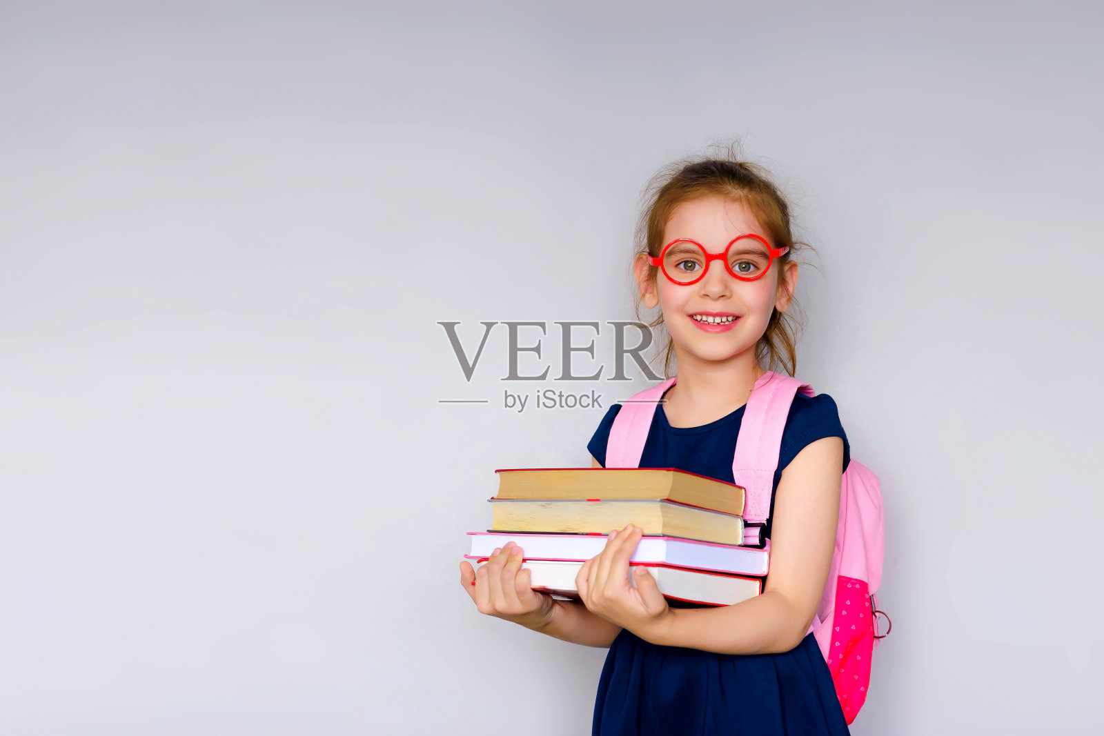 回到学校。一个戴着红眼镜拿着书包和书的金发女学生的肖像。灰色工作室背景。教育。对着镜头微笑。Copyspace。照片摄影图片