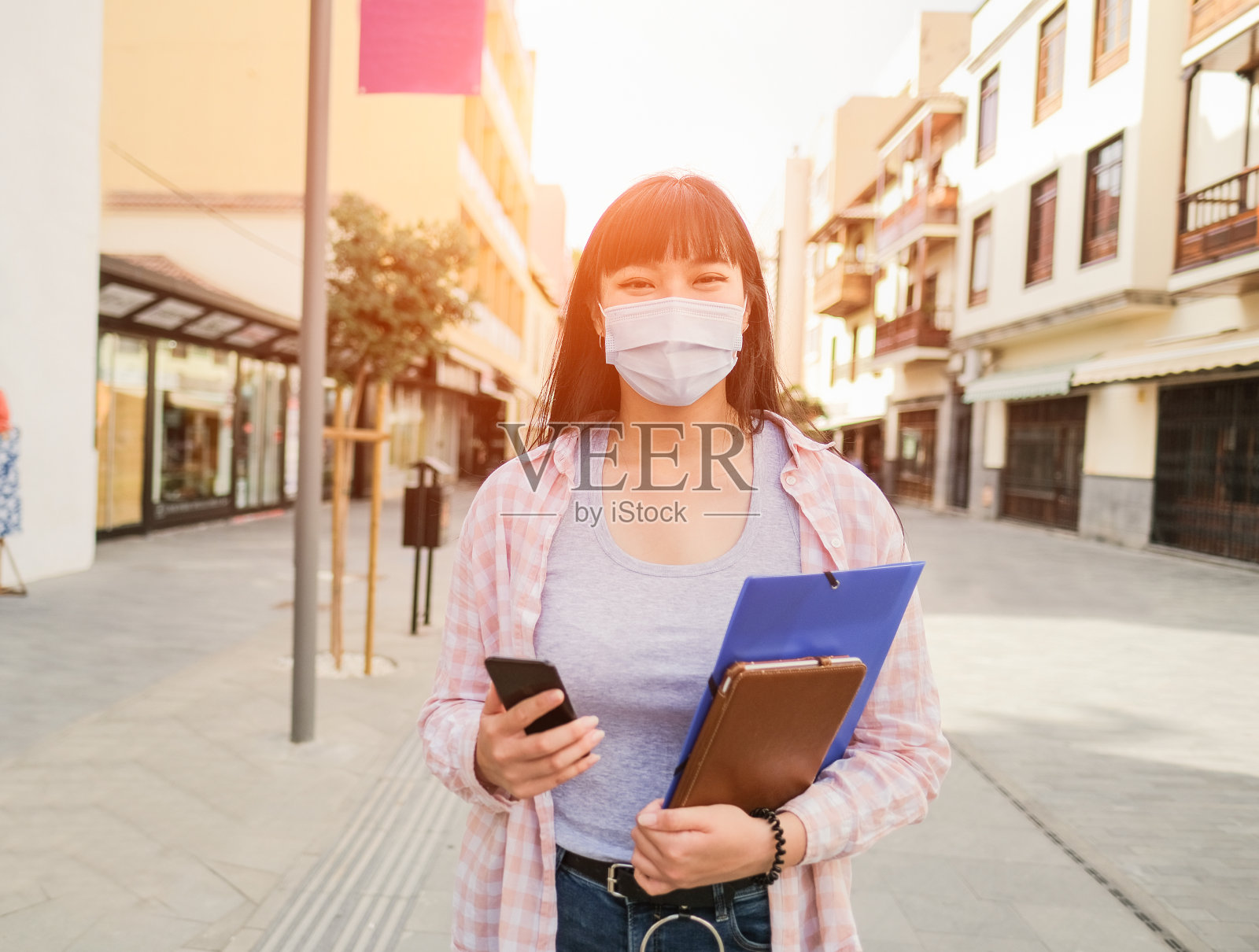 年轻的亚洲女性在城市中使用智能手机、抄写本和平板电脑，同时戴着外科口罩——冠状病毒生活方式照片摄影图片