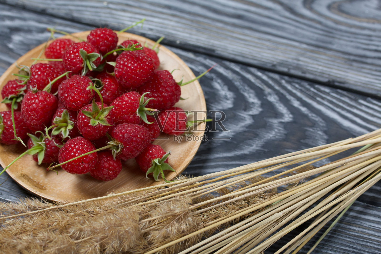 带尾巴的覆盆子躺在木托盘上。附近是一束带小穗的干草。在黑色的黑板上，具有富有表现力的木质纹理。照片摄影图片