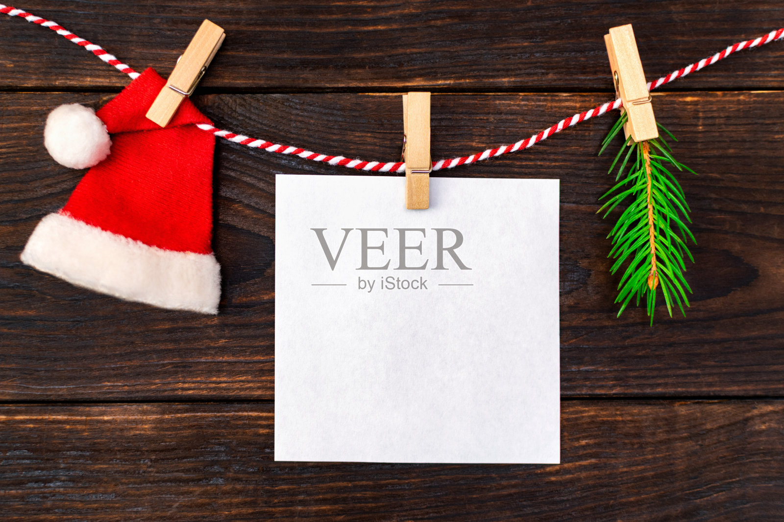 圣诞老人的红帽子，圣诞树的树枝和白纸空白的便条或信息挂在绳子上，并附上一个晾衣夹。木制背景上的传统新年标志。照片摄影图片