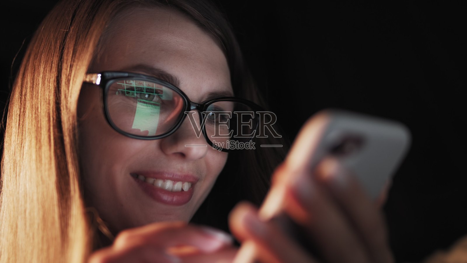 美丽的年轻女子躺在床上，使用智能手机和睡前微笑。一个女人晚上在床上用手机。迷人的女孩看手机。照片摄影图片