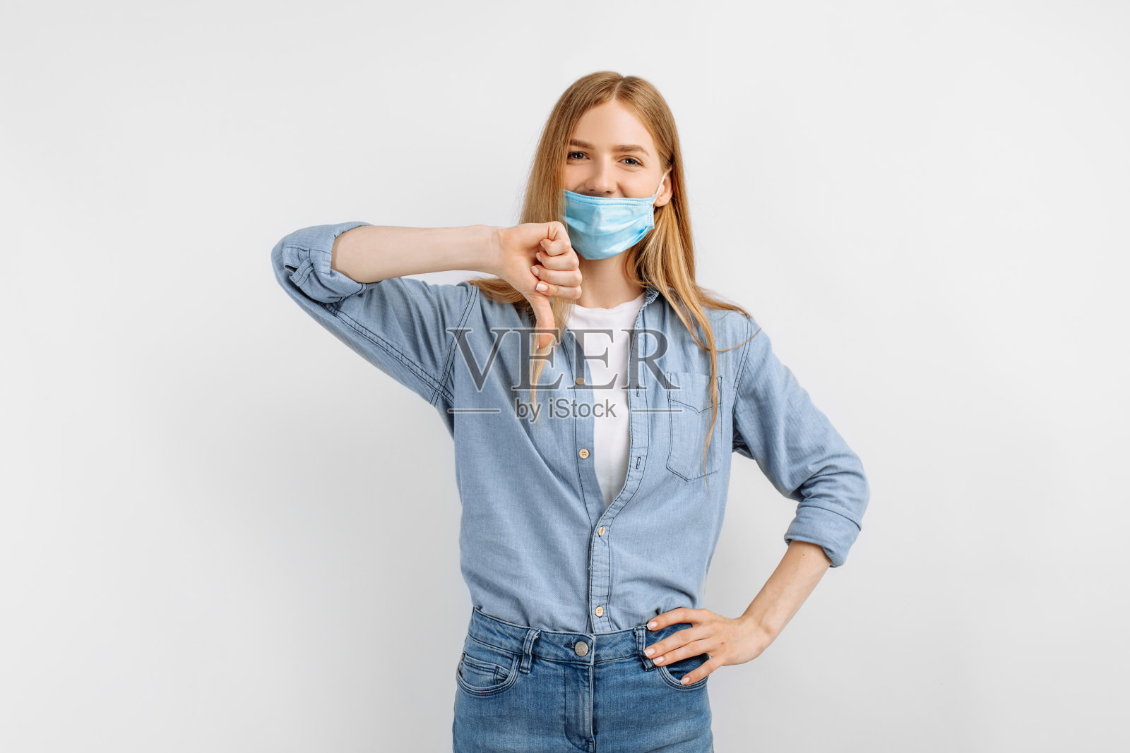 一个脸上戴着医用防护面具的不满的年轻女子，在白色背景下做出了一个拇指向下的手势，这是一个不友好的手势照片摄影图片