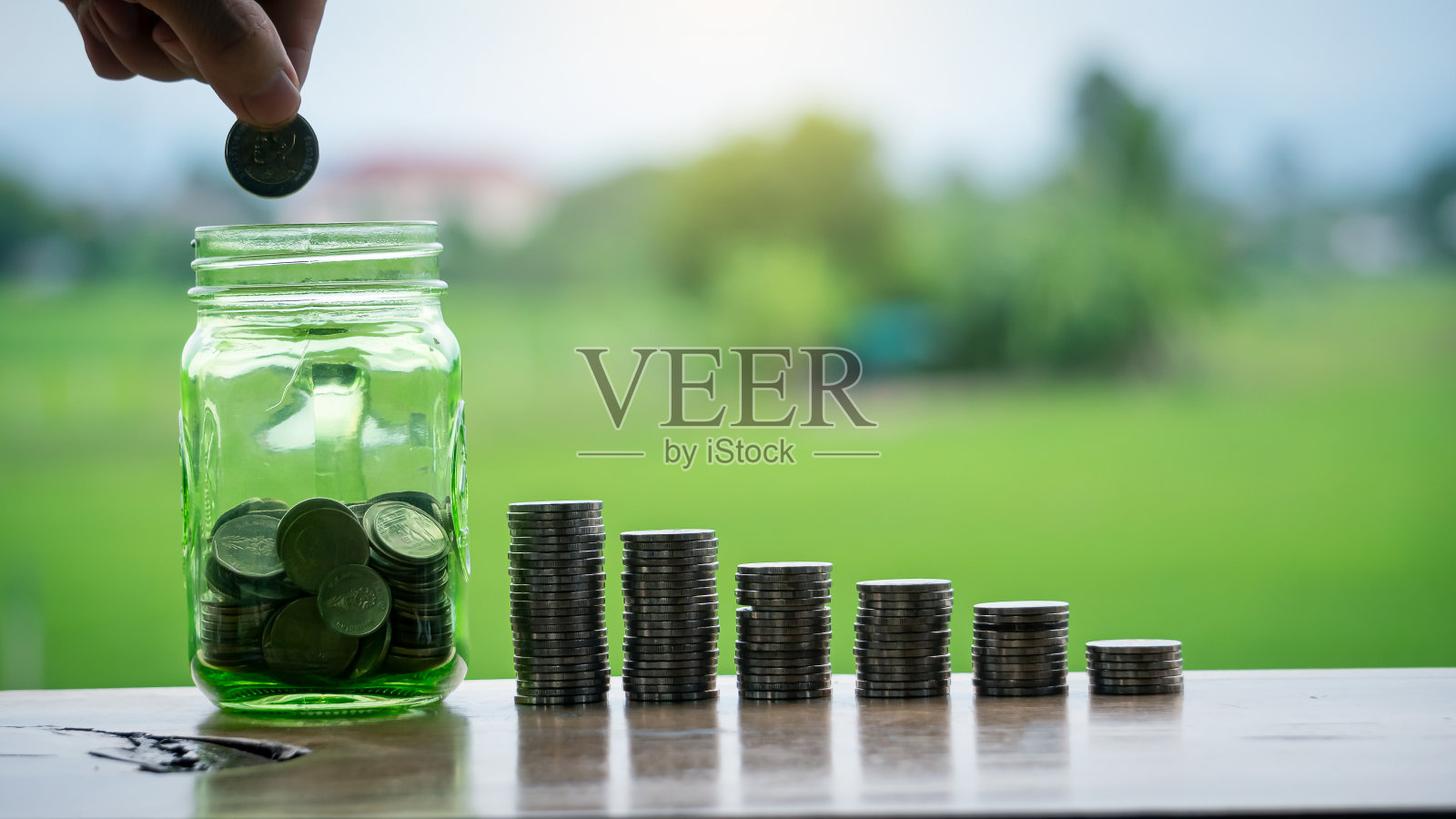 一只手拿着一枚硬币，把存钱罐和一堆硬币排成一排，就像一张图在绿色背景上模糊了存钱的概念照片摄影图片