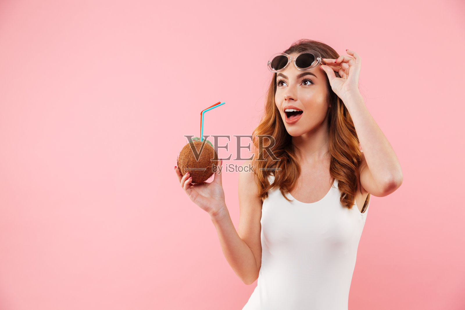 迷人的黑发女人在泳衣和太阳镜微笑和看一边，而手持椰子饮料孤立在粉红色的背景照片摄影图片