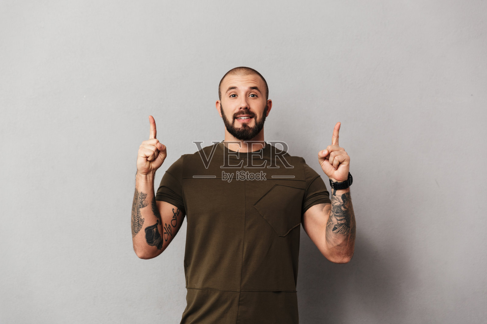 这是一幅没有刮胡子的快乐男子，双手纹身，在镜头前摆姿势，手指向上指着文案空间，孤立在灰色背景中照片摄影图片