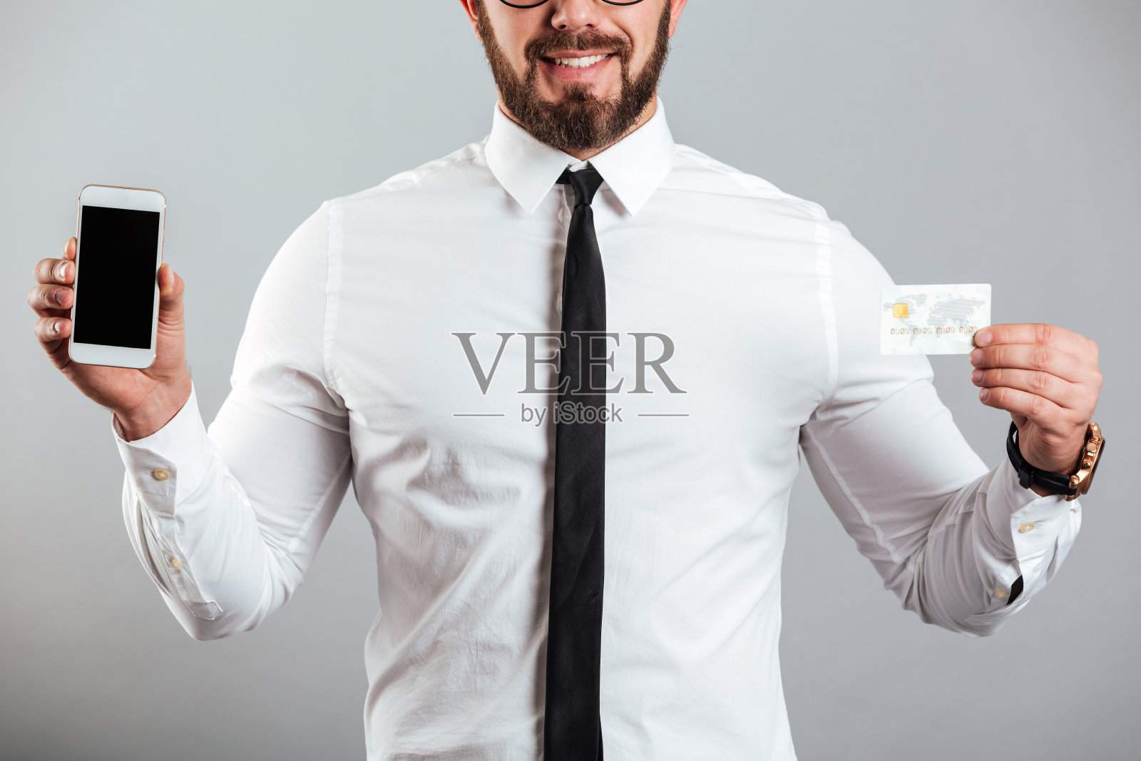照片中，一个穿着衬衫和领带、看上去像商人的男人，展示着智能手机和信用卡，孤立在灰色背景中照片摄影图片