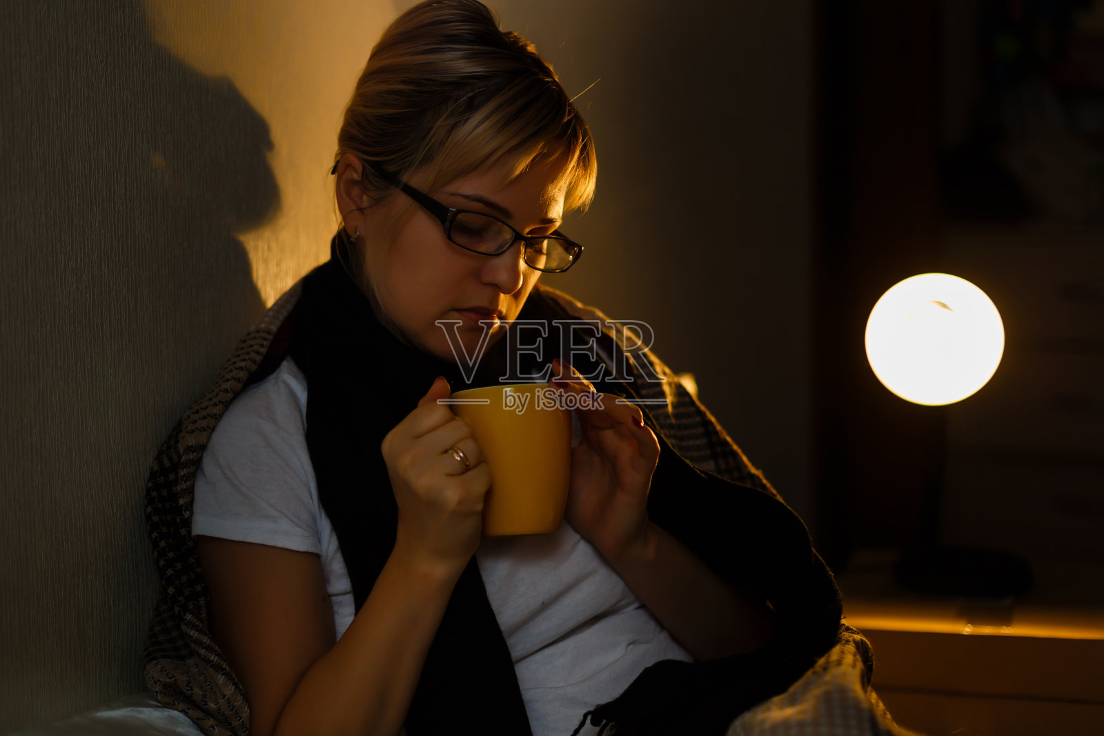 生病的女人，给你一杯茶。年轻沮丧的生病妇女在针织围巾拿着一杯茶躺在床上的特写图像。感觉不好。照片摄影图片