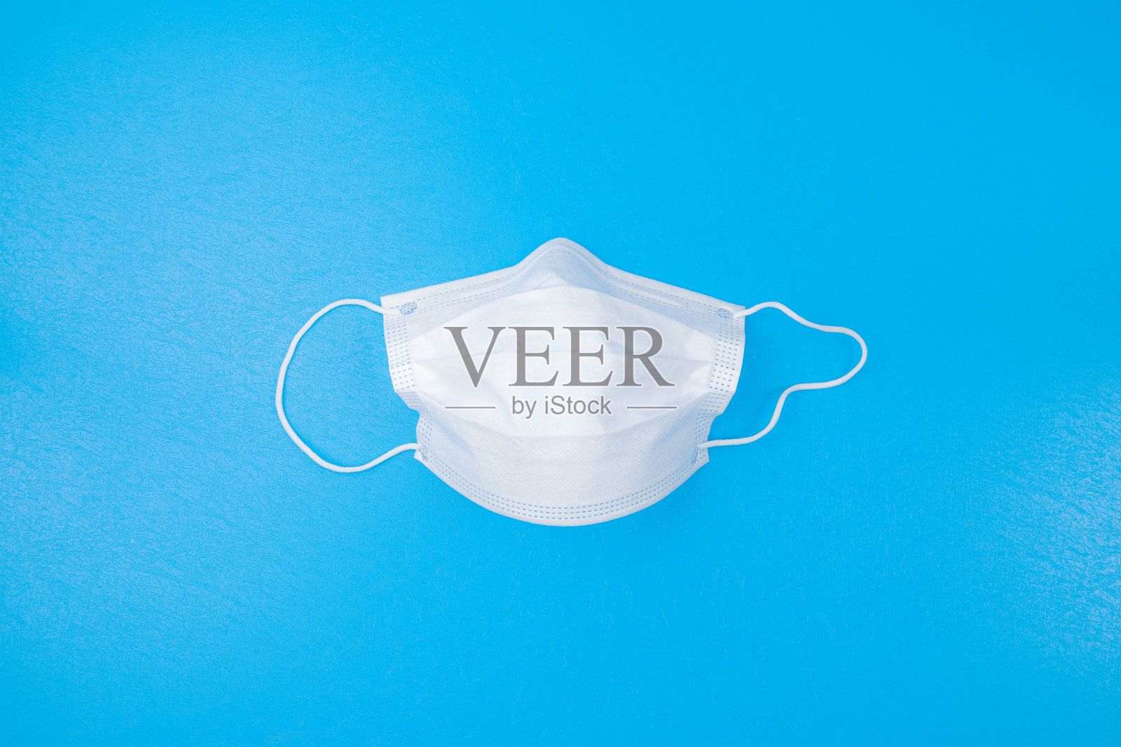 一次性医用口罩，蓝色背景，白色纸眼。带着医用口罩微笑切断脸部。阻止冠状病毒传播，自卫。复制照片摄影图片