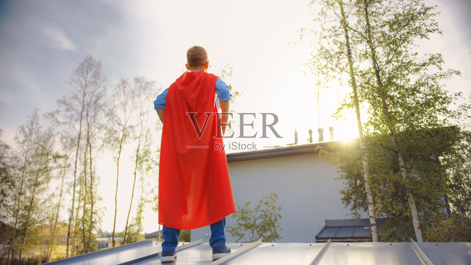 男孩正在扮演一个超级英雄的角色。他站在屋顶上，双手放在腰上。年轻人穿着一件鲜红色的斗篷。他在看太阳。照片摄影图片