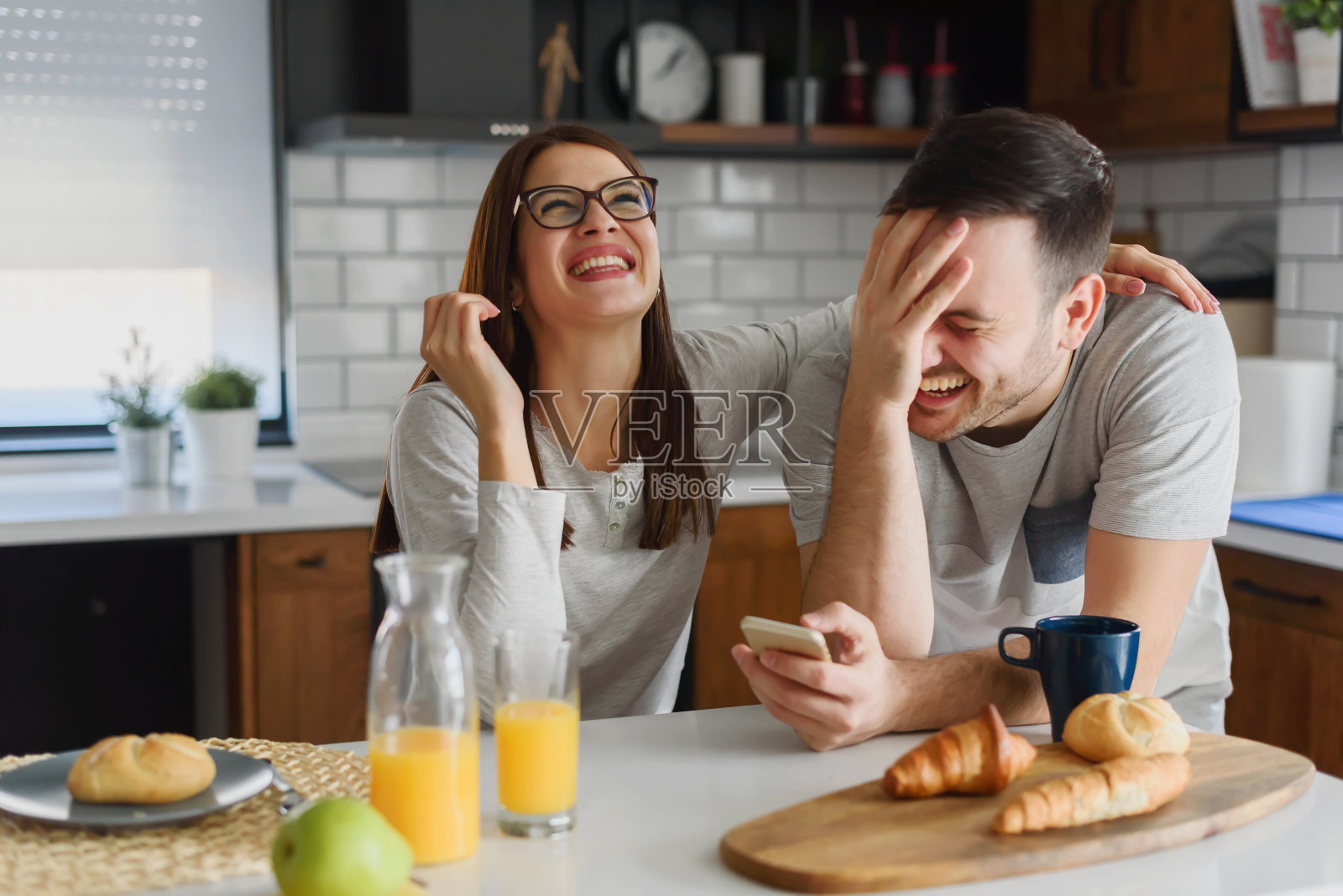 微笑的夫妇在家里厨房享受美丽的早晨，吃早餐，喝咖啡和果汁，看智能手机照片摄影图片