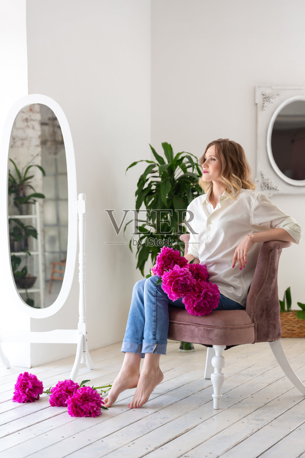 一个穿着白色衬衫和蓝色牛仔裤的女人，手里拿着粉红色的牡丹花，坐在一个白色的房间的椅子上照片摄影图片