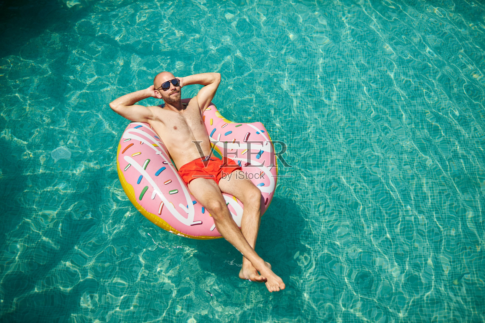 一名训练有素的中年男子，穿着红色泳裤，戴着墨镜，躺在充气甜甜圈里，享受着假期。照片摄影图片