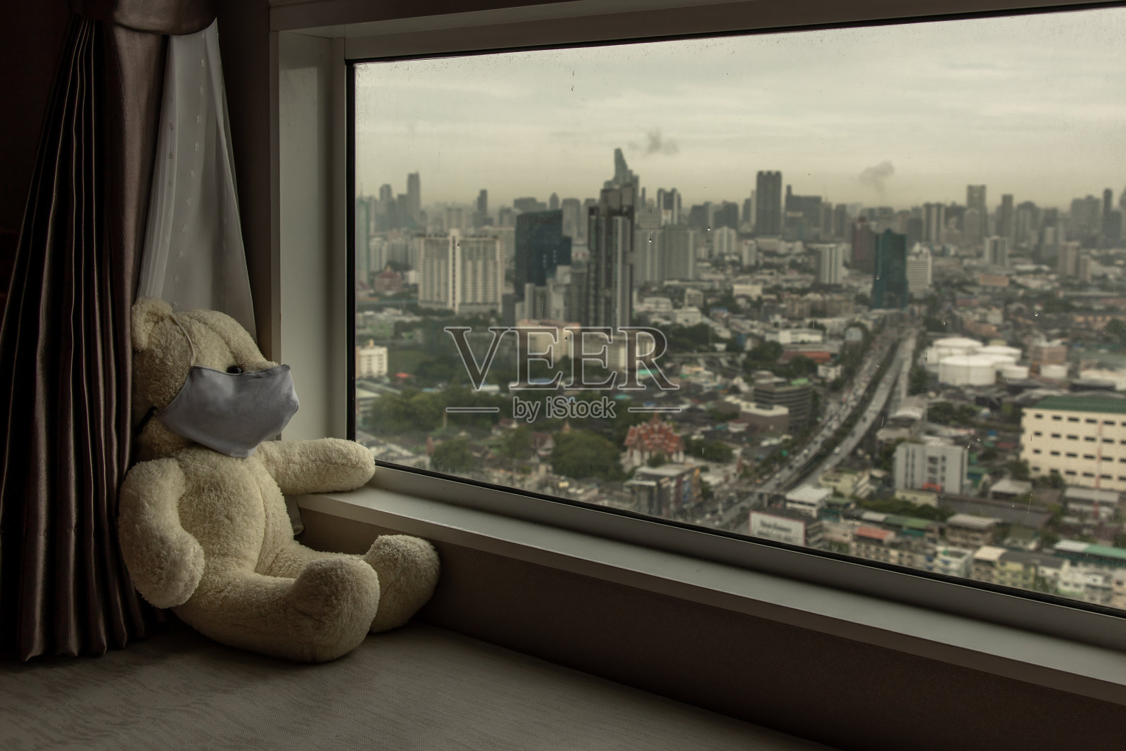 悲伤的泰迪熊戴着医用防护口罩坐在窗台上望着窗外。照片摄影图片