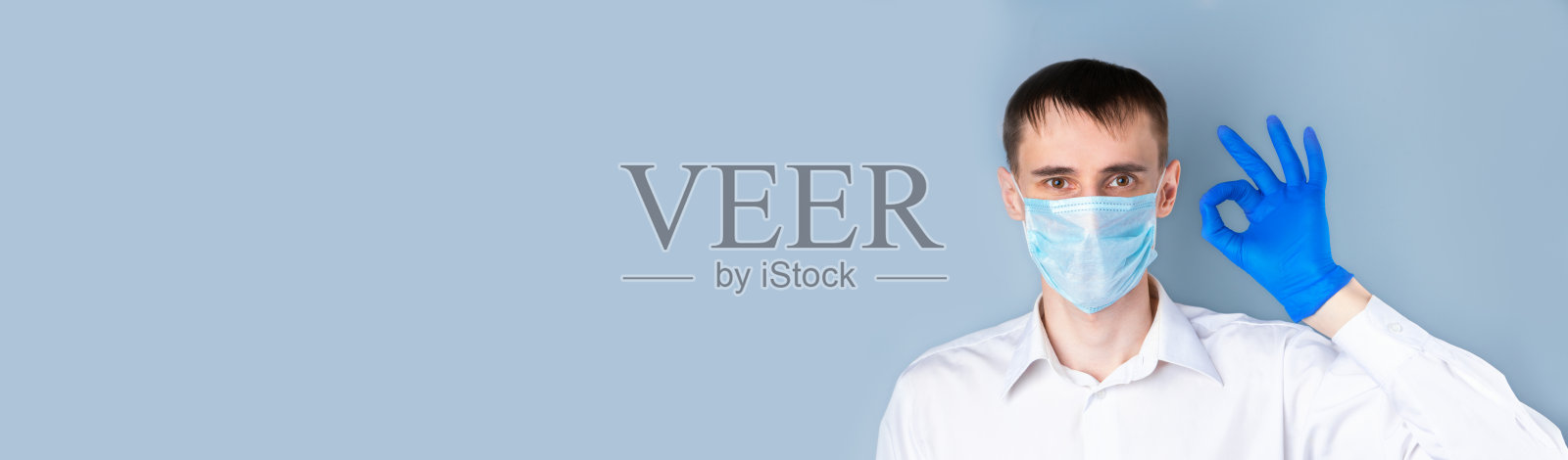 一名戴着医用口罩的男子戴着蓝色医用手套，打着“OK”的手势，身穿白衬衫，背景为灰色，文字空白。横幅。冠状病毒,Covid-19照片摄影图片
