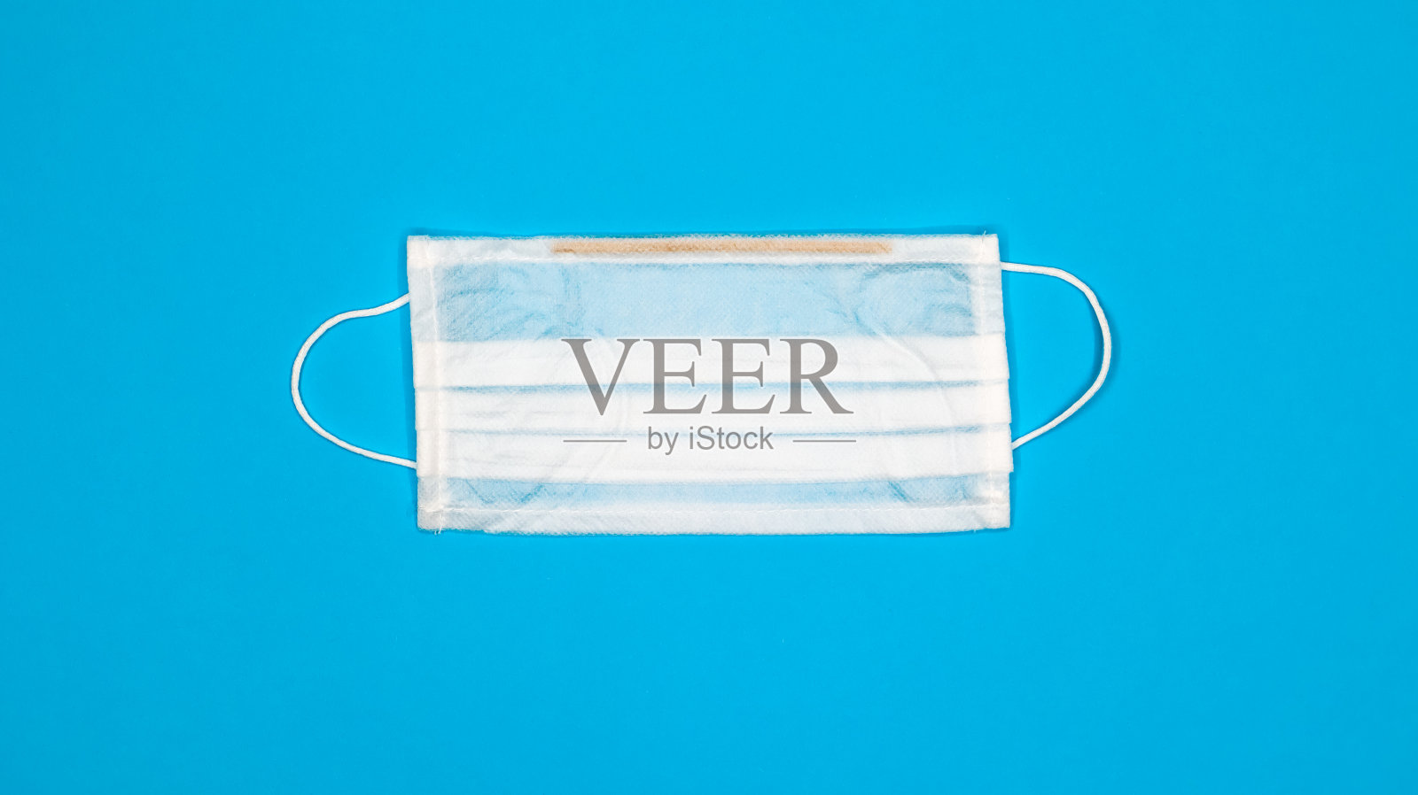 一个三层的一次性外科口罩，用橡胶夹子盖住嘴和鼻子，背景为蓝色。保护细菌，保健和医学的概念。新型冠状病毒肺炎一个危险的病毒照片摄影图片