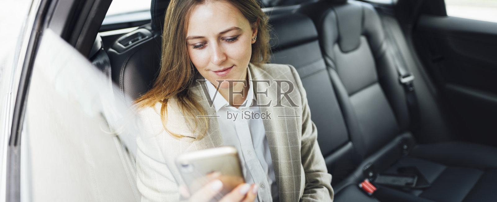 广角镜头。美丽的商务女性在出租车上使用智能手机。年轻的女商人坐在车后座上打电话照片摄影图片
