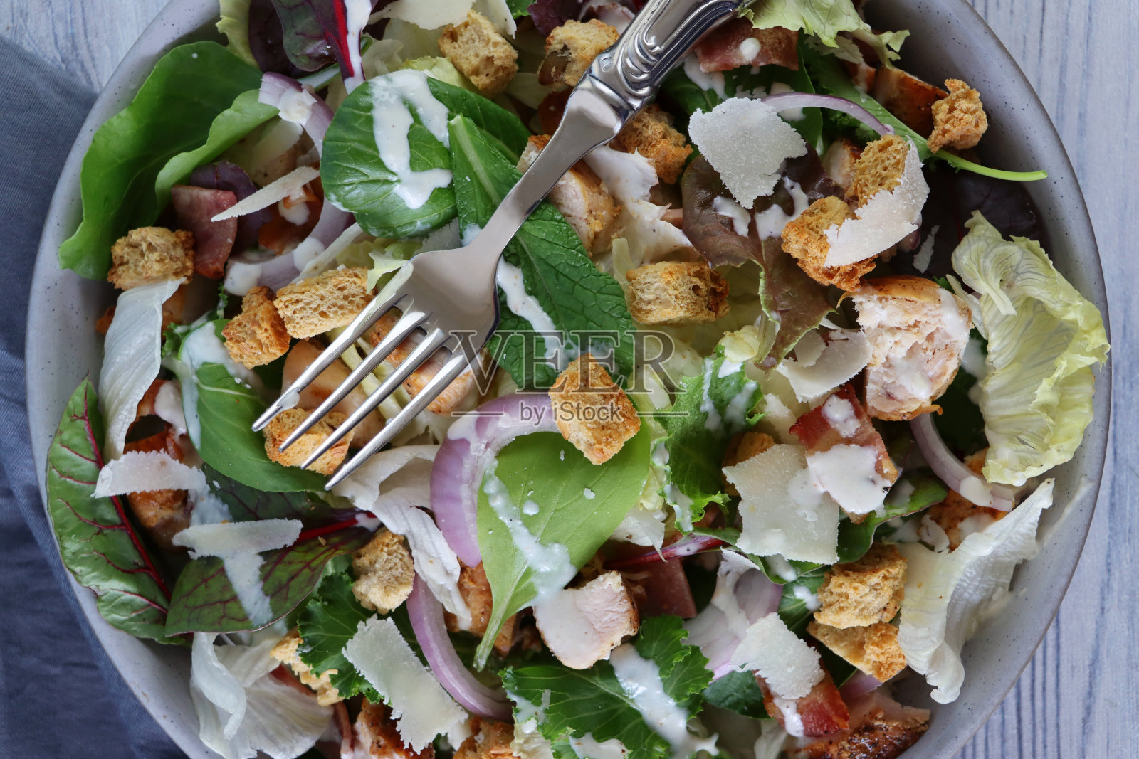 特写的鸡肉凯撒沙拉在碗里，生菜，红洋葱，甜菜根叶，培根丁，烤鸡，上面放着面包丁和帕尔玛干酪，除了灰色的茶毛巾，还有金属叉，木纹背景，俯瞰照片摄影图片