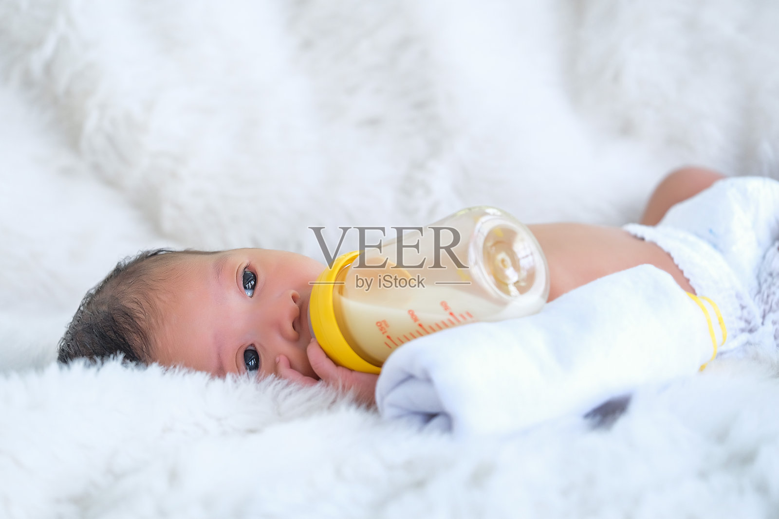 白色的舞会床上的婴儿正在吸奶瓶里的牛奶。照片摄影图片