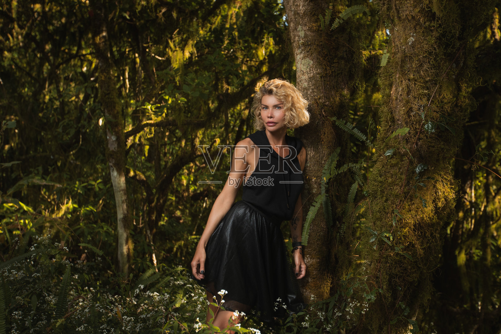 美丽的金发美女站在森林的一棵树旁，穿着黑色的裙子，戴着头巾照片摄影图片