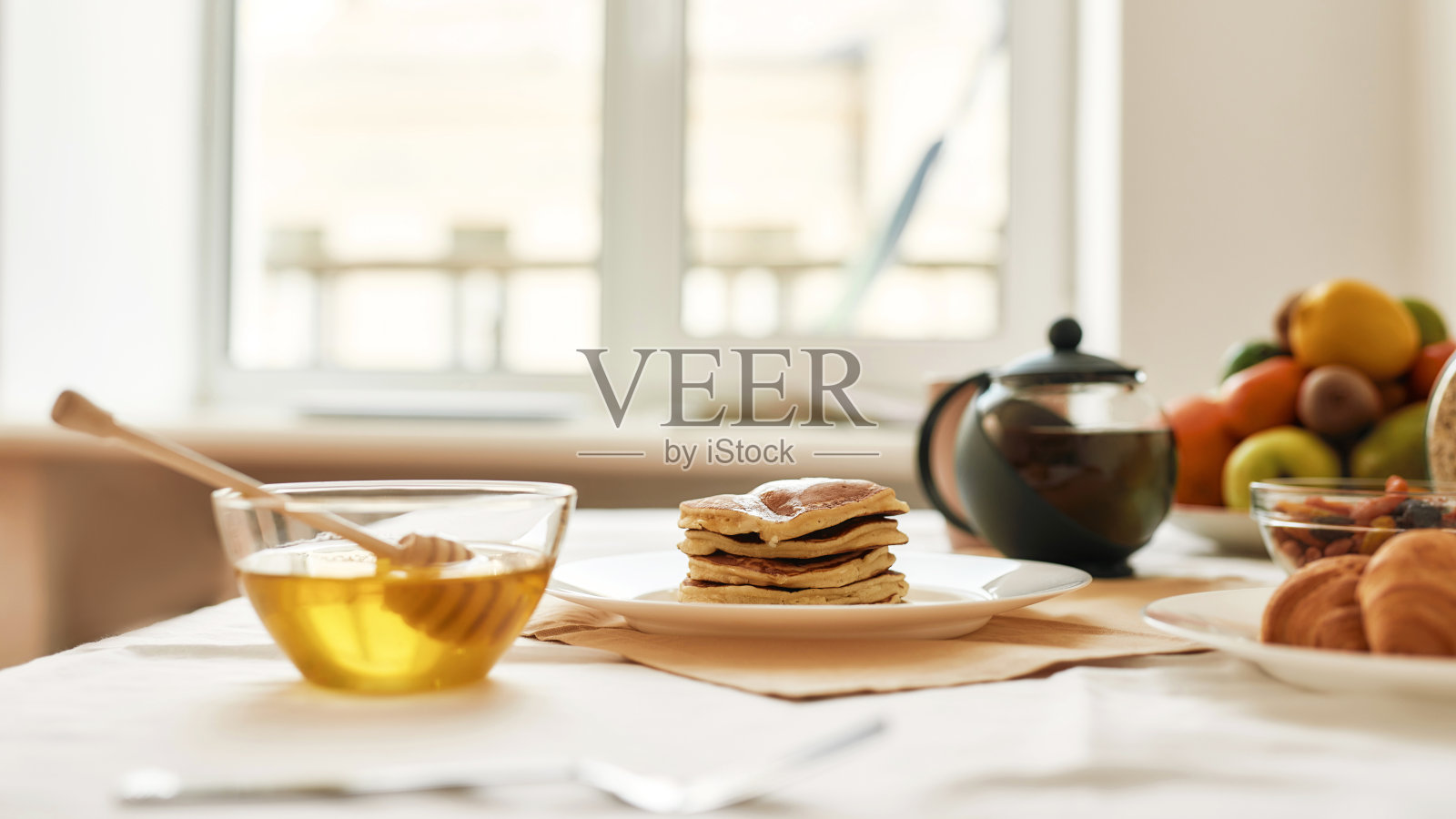 一堆香甜可口的煎饼，碗里放着蜂蜜，茶壶里放着牛角包和各种水果，早餐摆在桌上照片摄影图片