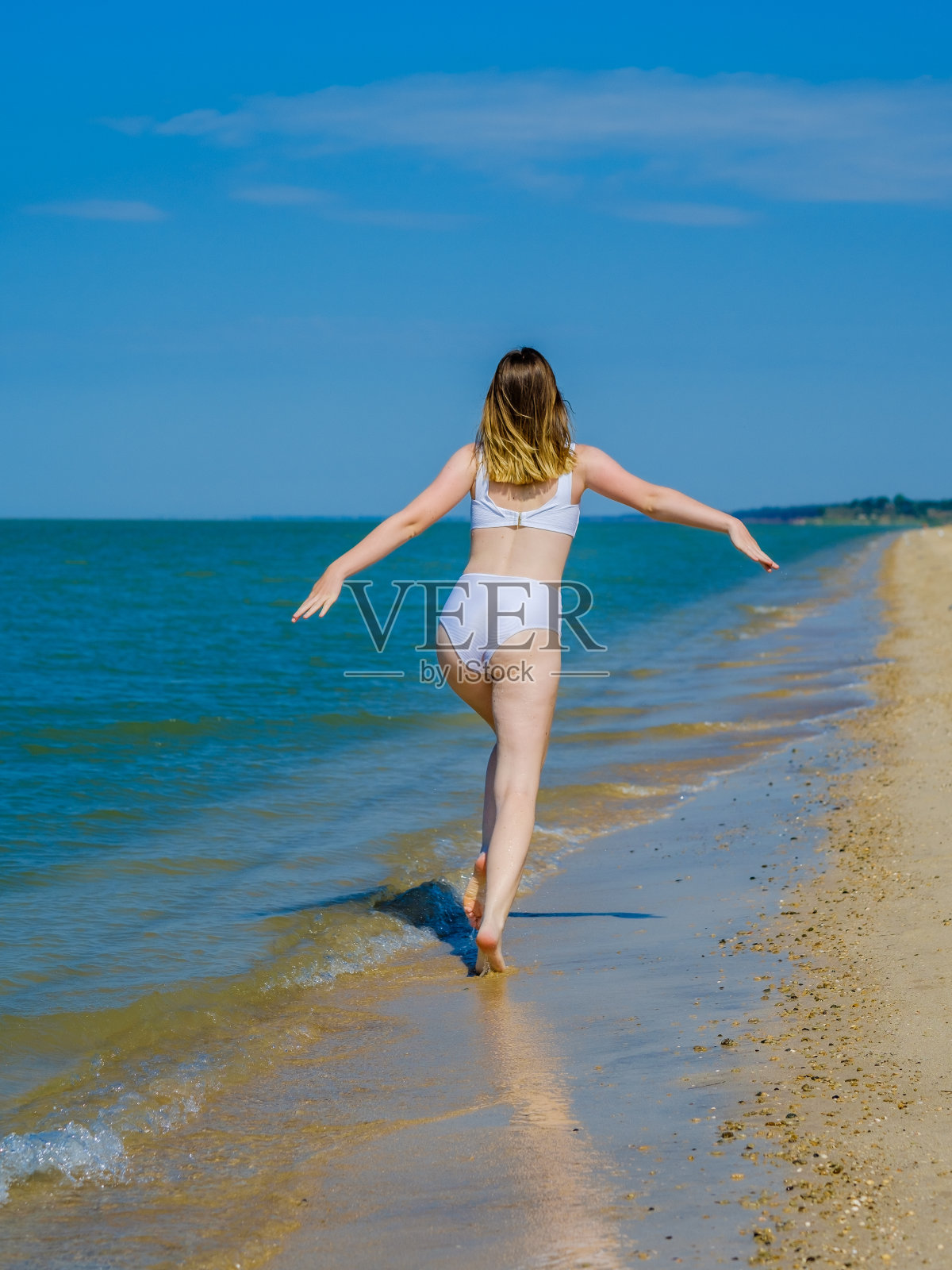 一个女孩的背影沿着沙滩跑向远方海水的飞溅。那个女孩穿着白色的泳衣，戴着太阳镜。自由。健康。夏天阳光灿烂的日子。本空间照片摄影图片