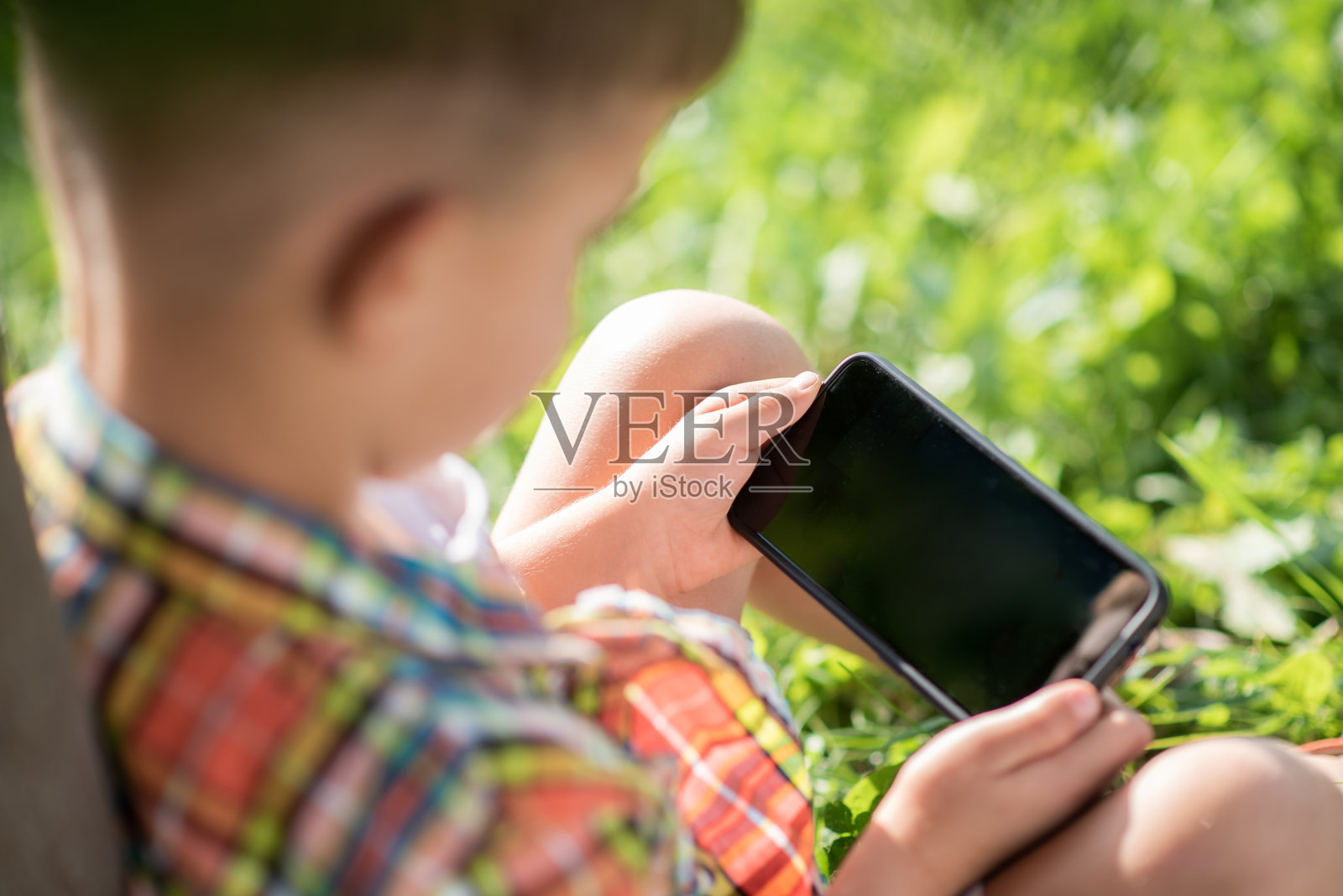 一个美丽的孩子坐在草地上看手机在夏天日落。手里拿着手机的可爱男孩照片摄影图片