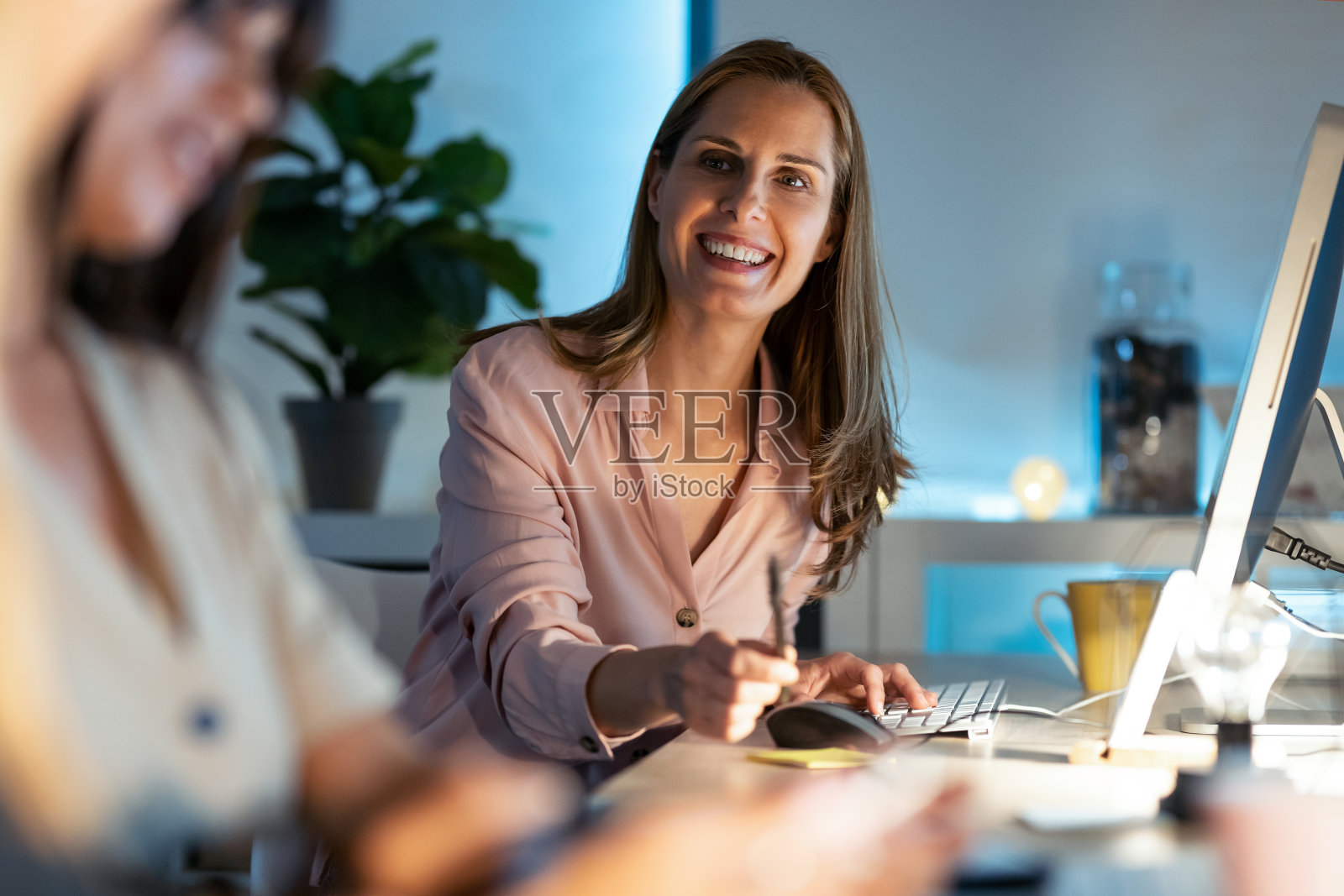 微笑着美丽成熟的商业女人与她的同事聊天，同时工作与电脑在办公室。照片摄影图片