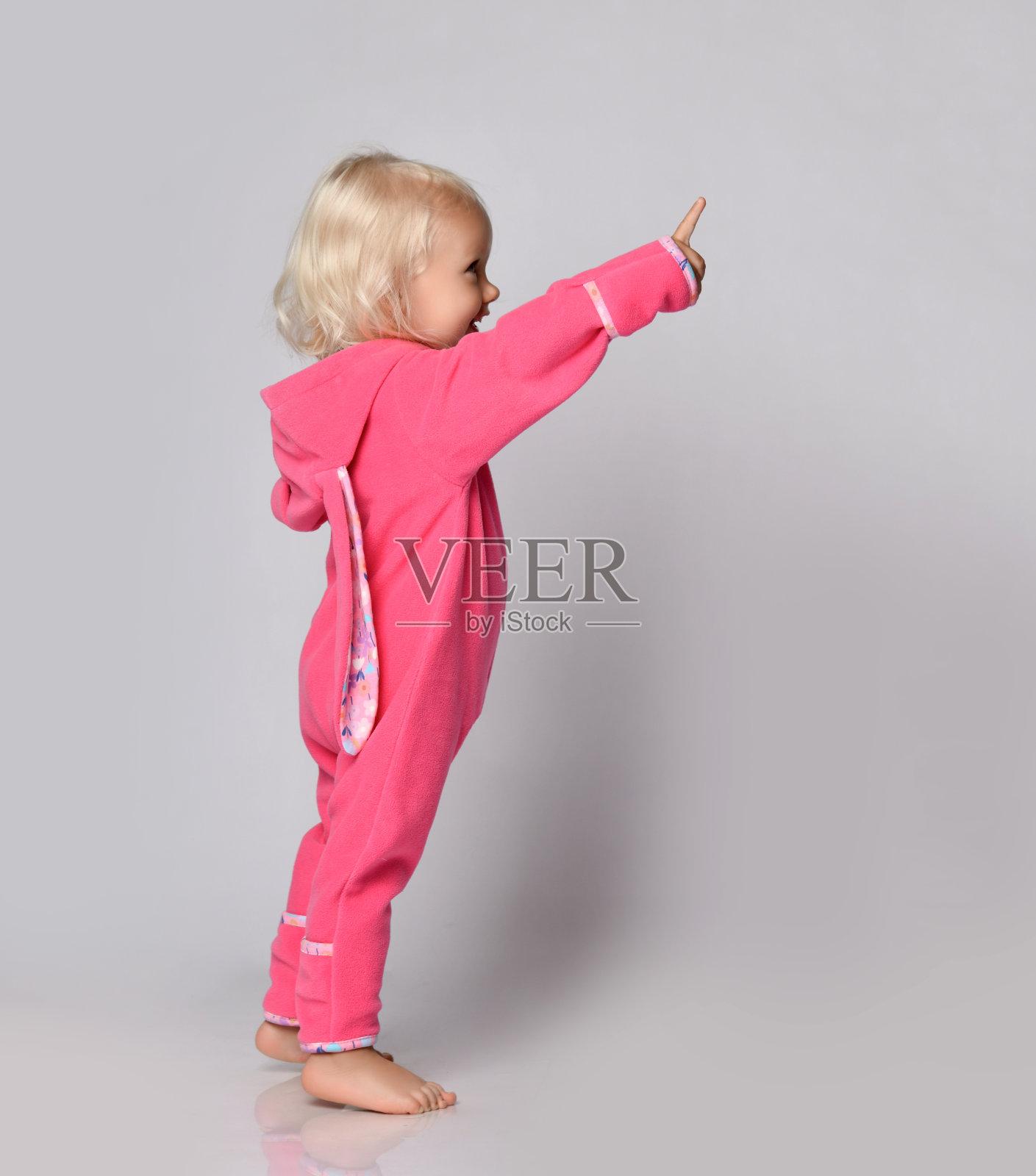 小可爱的金发积极的婴儿女孩在粉红色温暖舒适的连衣裤站着，用手指向上照片摄影图片