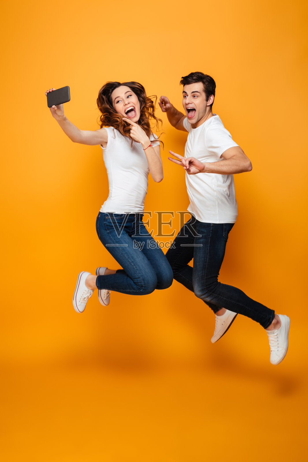 两名快乐的20多岁的男女一边用手机自拍，一边在黄色背景上跳来跳去，打着手势照片摄影图片