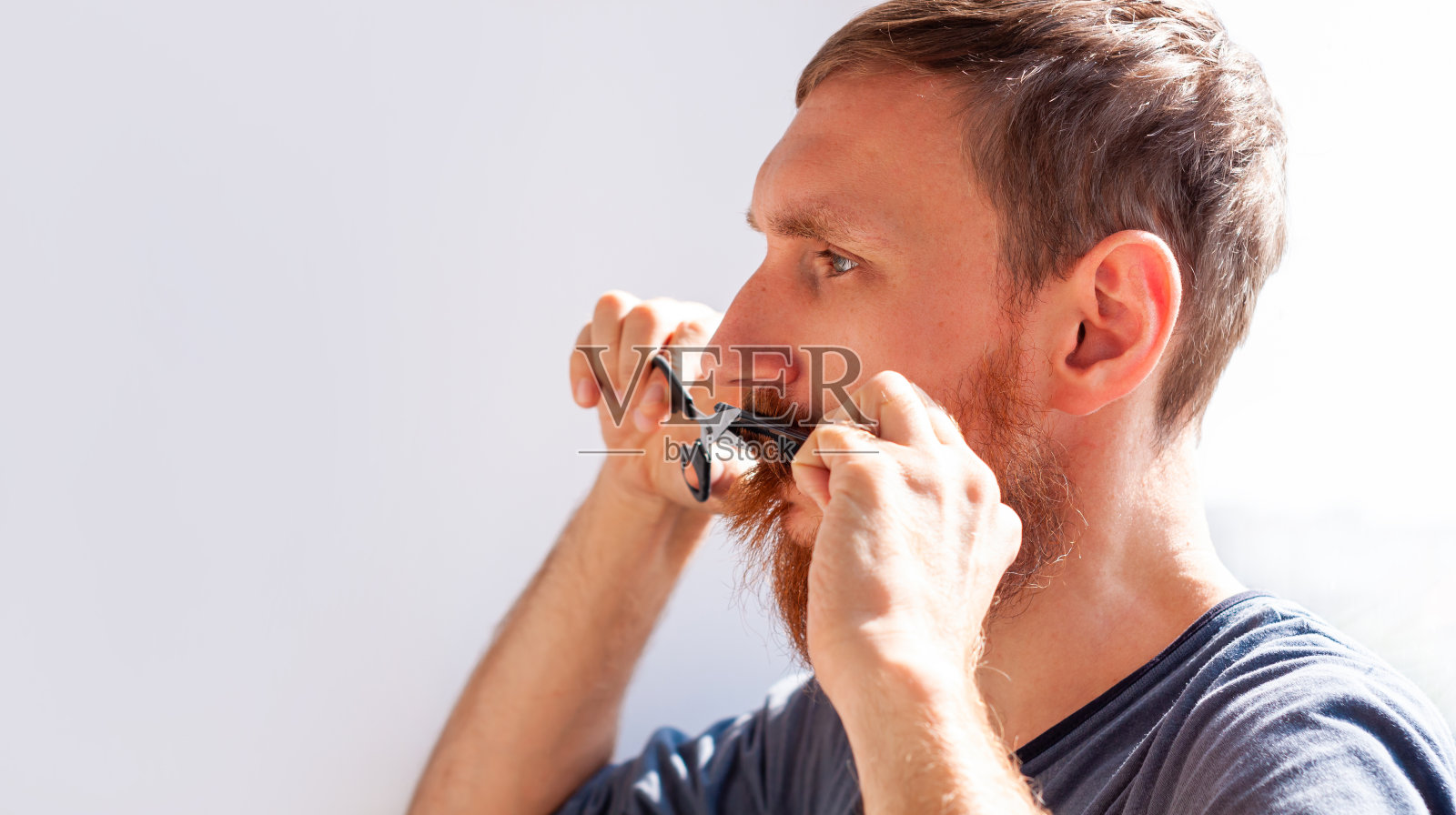 男人用剪刀和梳子剪自己的胡子和小胡子。白人红胡子男性在家修剪面部毛发。照片摄影图片