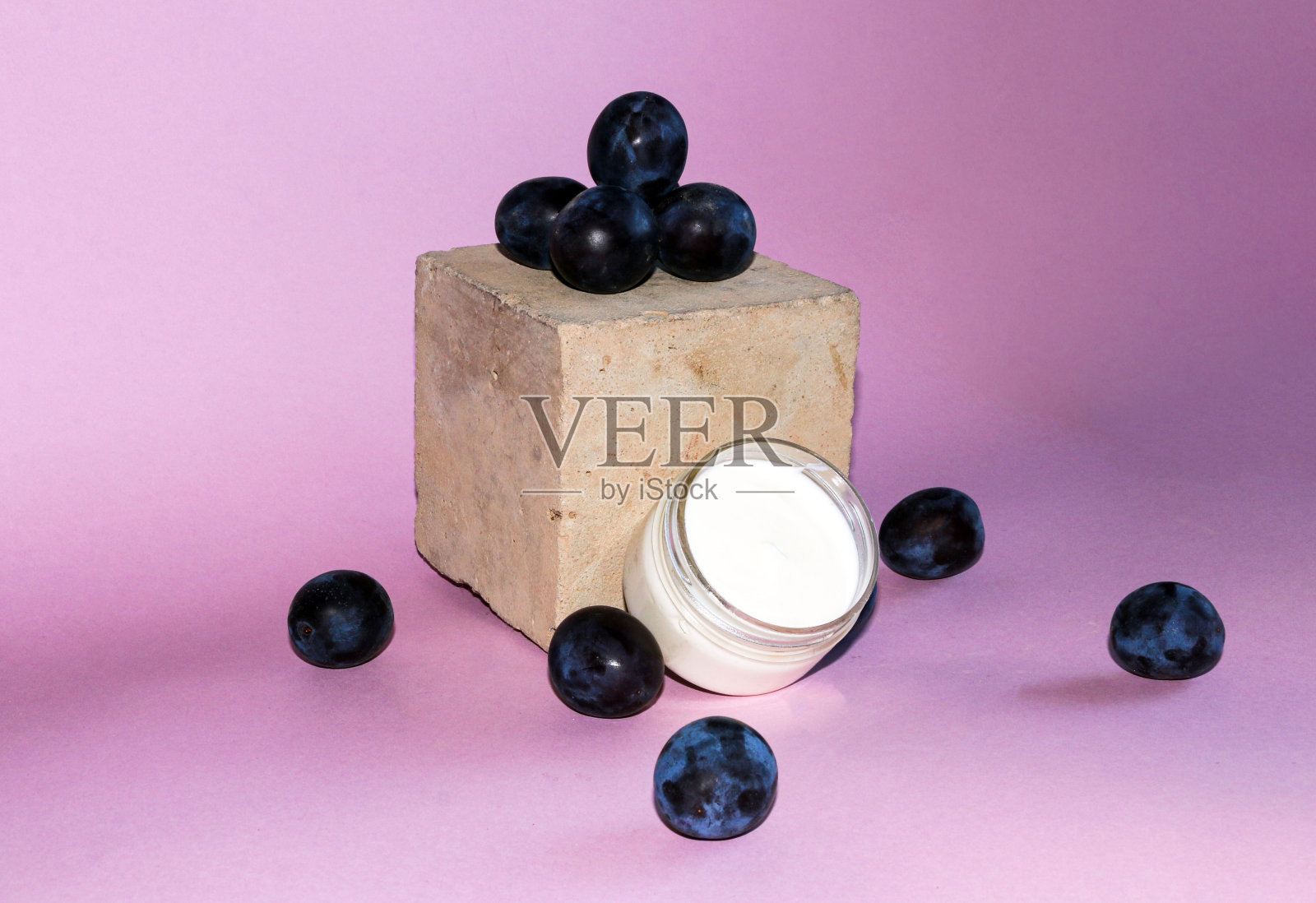化妆品组合与成熟的深蓝色李子，和一个开放的罐子，白色温和的保湿霜，在粉红色的背景。照片摄影图片