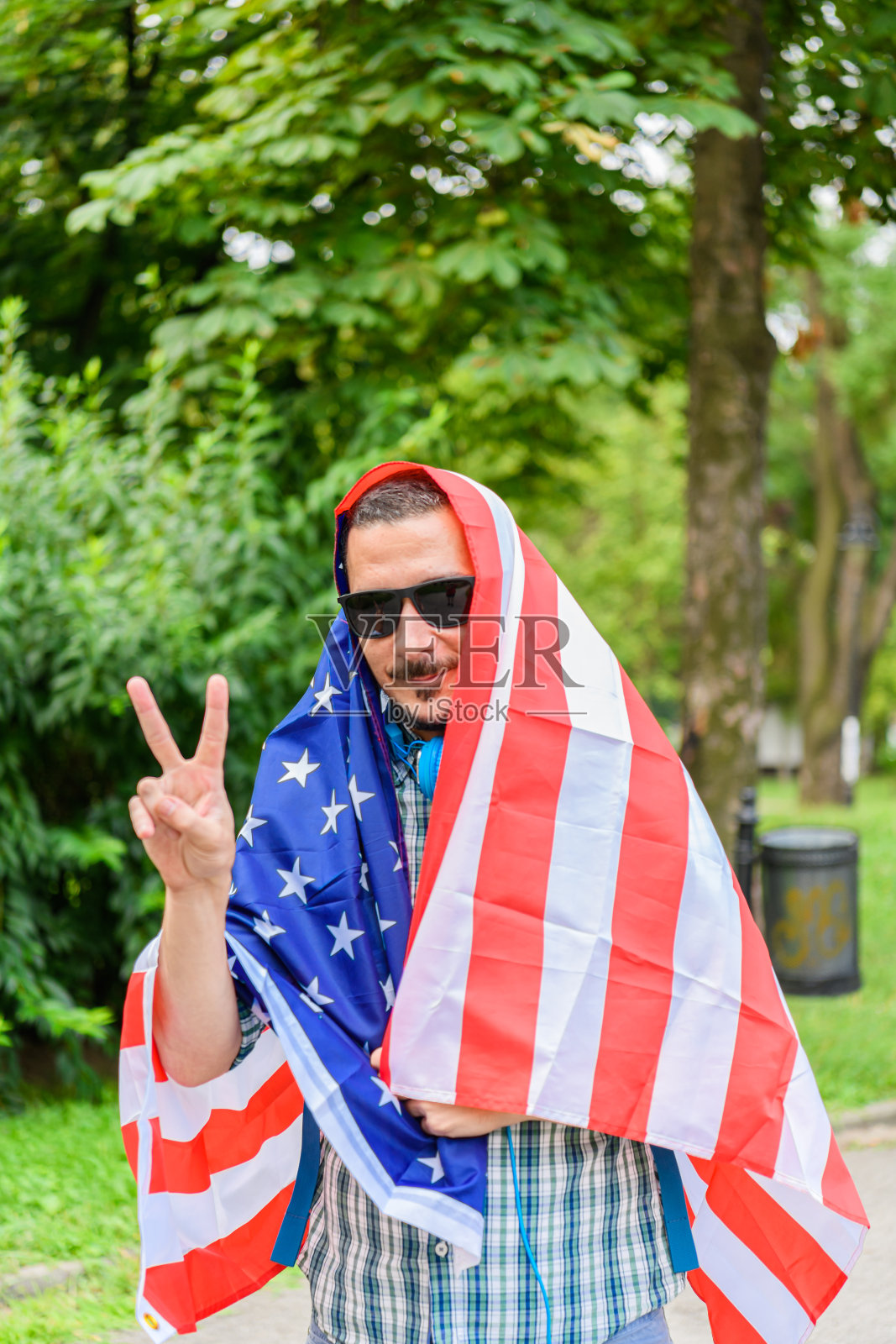 一个男人的肖像与背包在格子衬衫谁是走在公园和美国国旗显示一个V-Sign。照片摄影图片