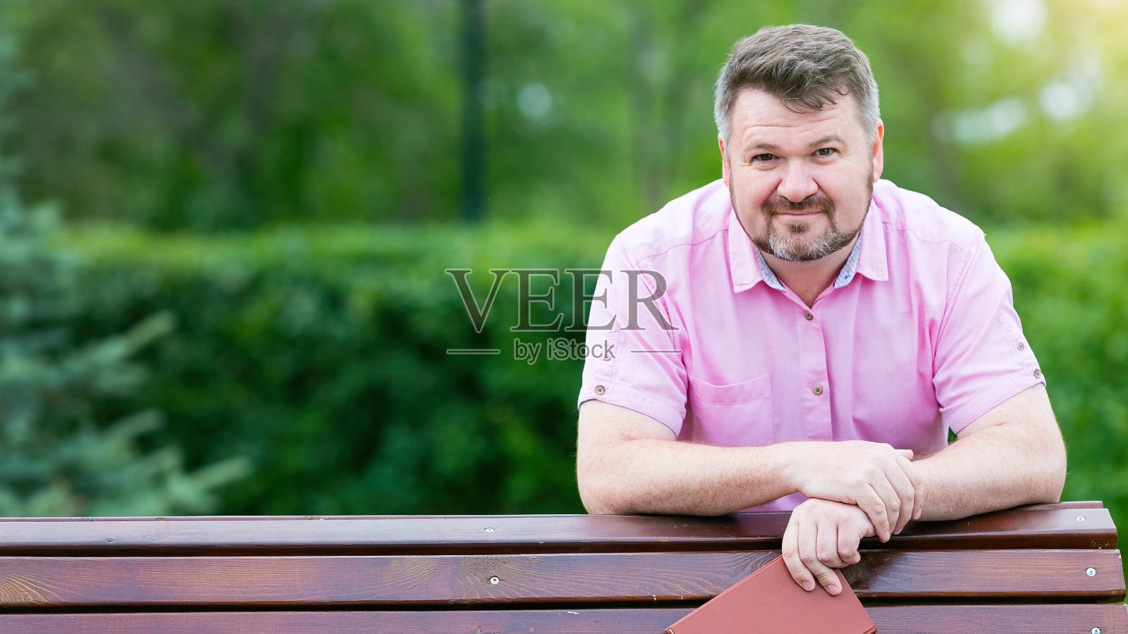 在公园里，一个穿着红衬衫留着胡子的成年男子靠在长凳上。他手里拿着一本书照片摄影图片