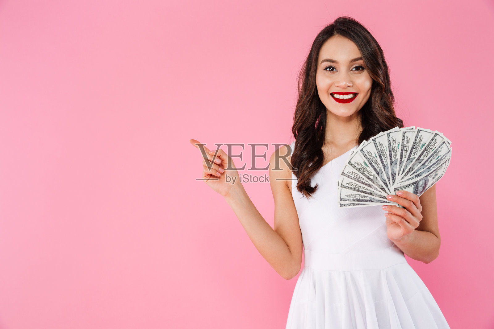 这是一个20多岁的成功亚洲女性的形象，她拿着一堆现金，微笑着把手指放在一旁，在粉红色的背景下孤立无间照片摄影图片