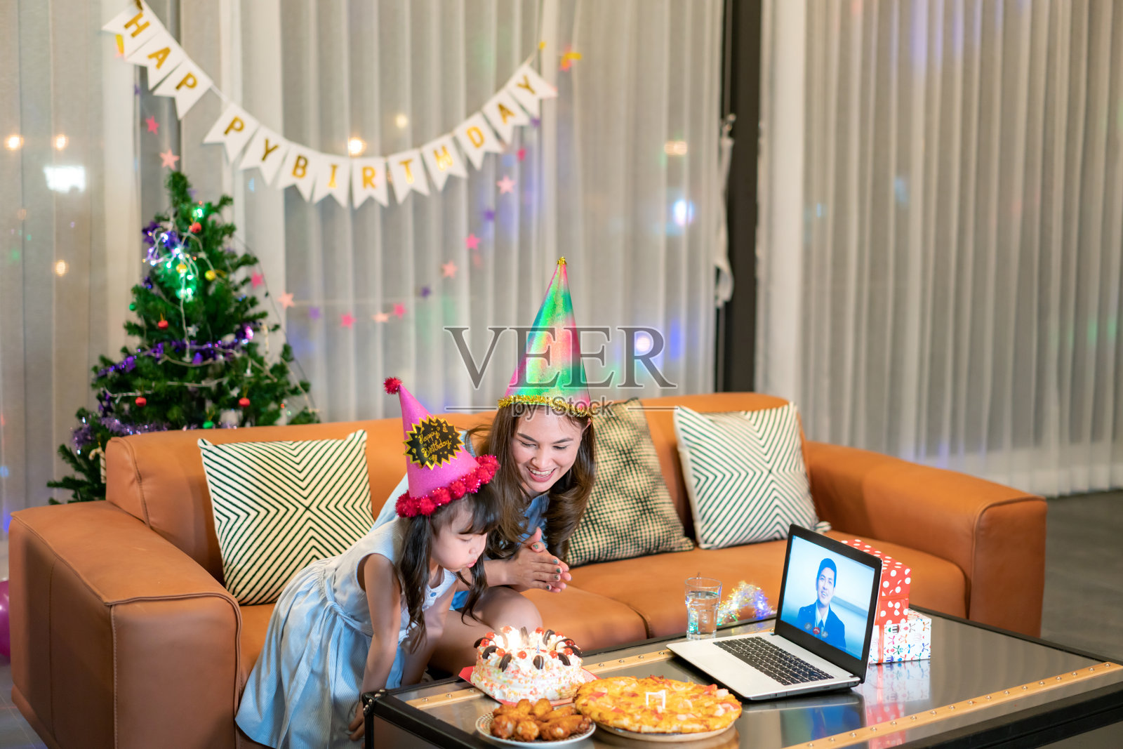 快乐的小女孩和妈妈在家庆祝生日，爸爸通过笔记本电脑视频通话，吹灭桌上的蛋糕蜡烛照片摄影图片