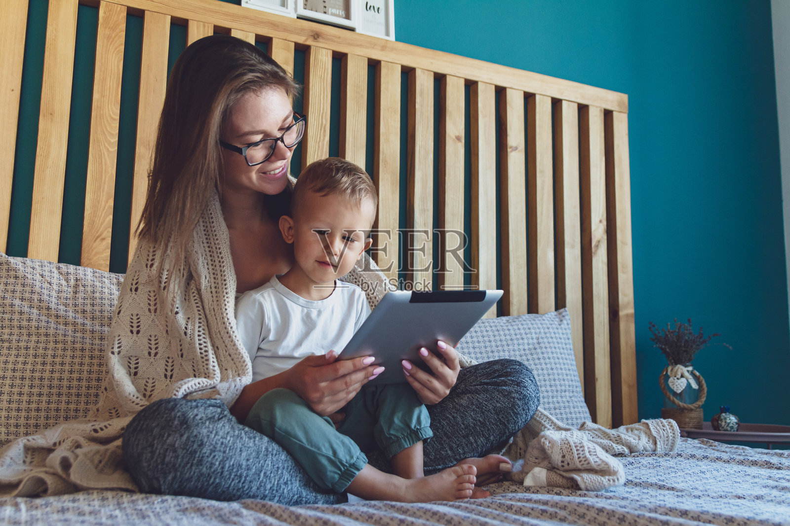 年轻的妈妈和儿子在床上舒适的毯子下面放着平板电脑照片摄影图片
