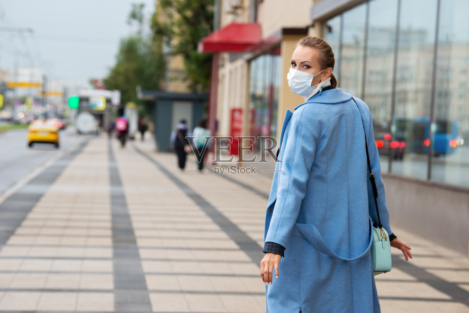 一个穿着蓝色外套，戴着医用防护口罩的女孩走在路边的人行道上。照片摄影图片