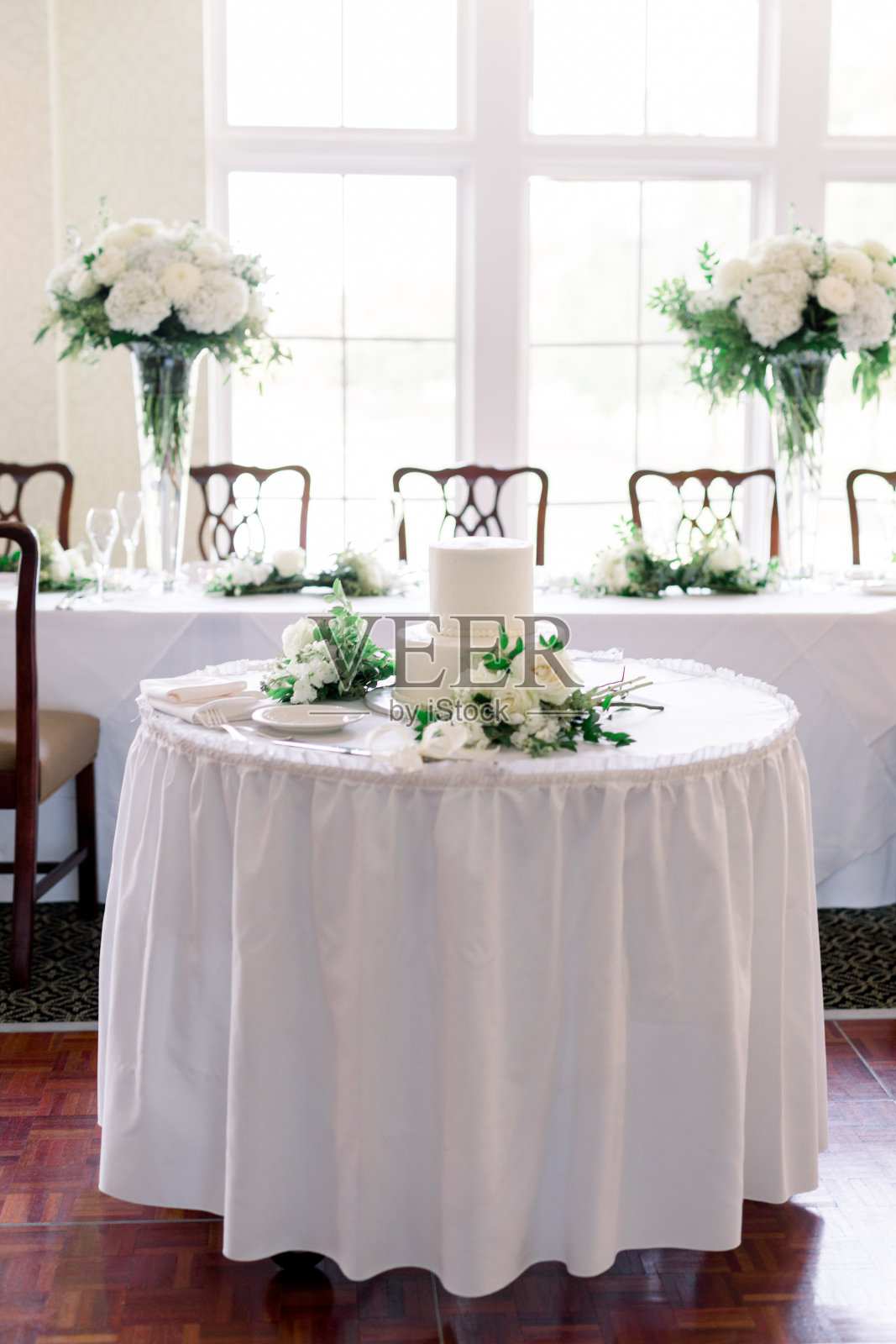 婚礼餐桌装饰与婚礼花束。在它的后面是一张长桌子，上面摆着华丽的椅子和两件大的鲜花，高高的花瓶里插着白色和绿色的花朵。照片摄影图片