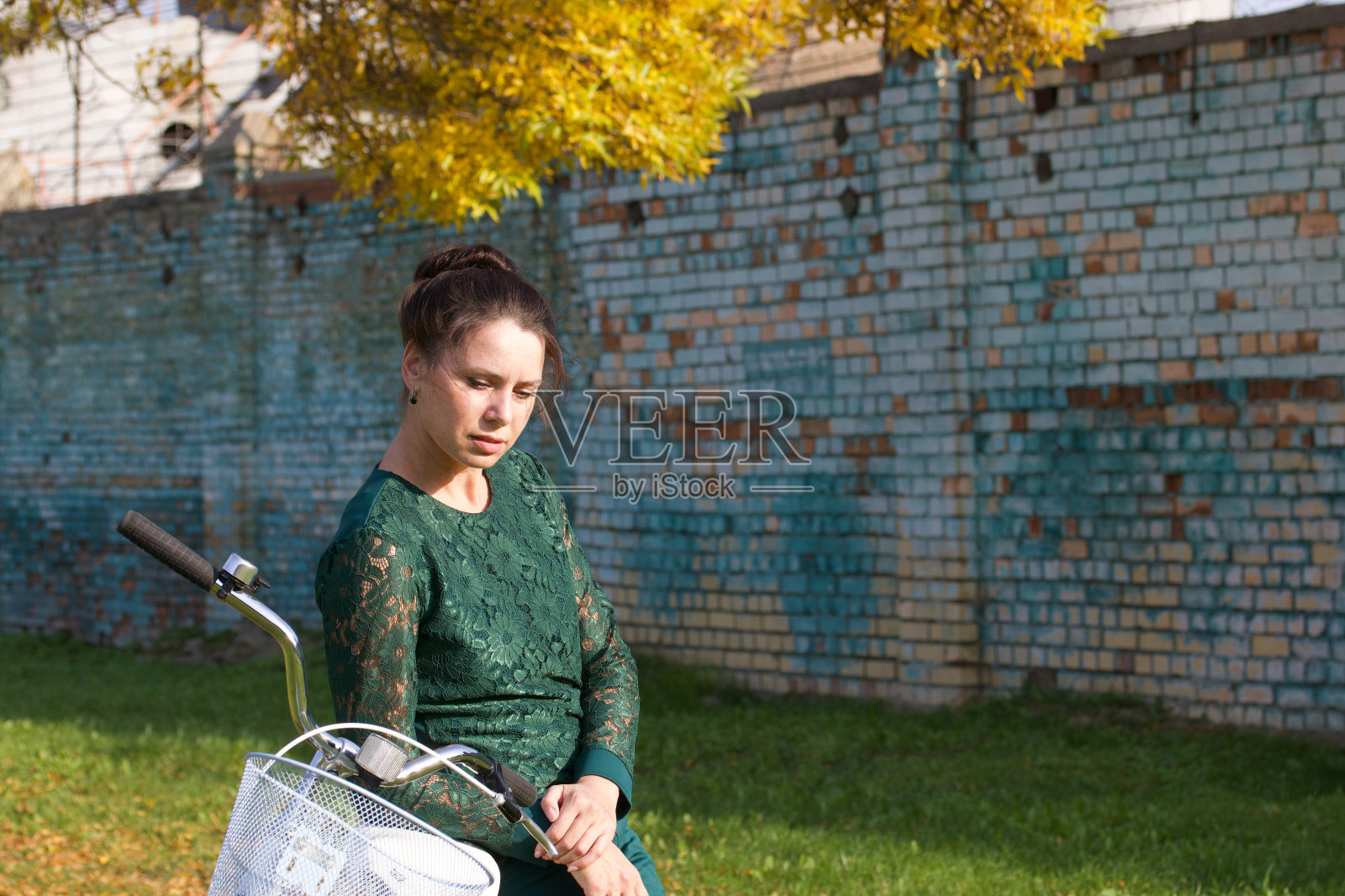 穿绿色运动服的女孩。站着，靠在自行车上。在黄叶飘落的小路上。以砖墙为背景。照片摄影图片