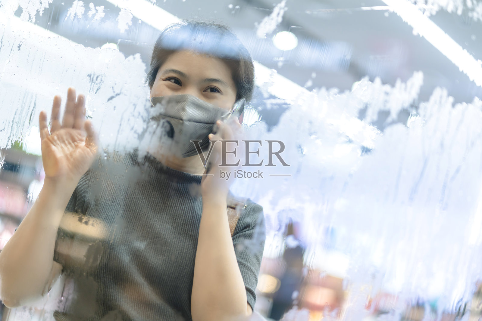 亚洲女性女性戴着防毒面具透过窗户看，用水珠雨滴手握智能手机交流新常态的生活方式照片摄影图片