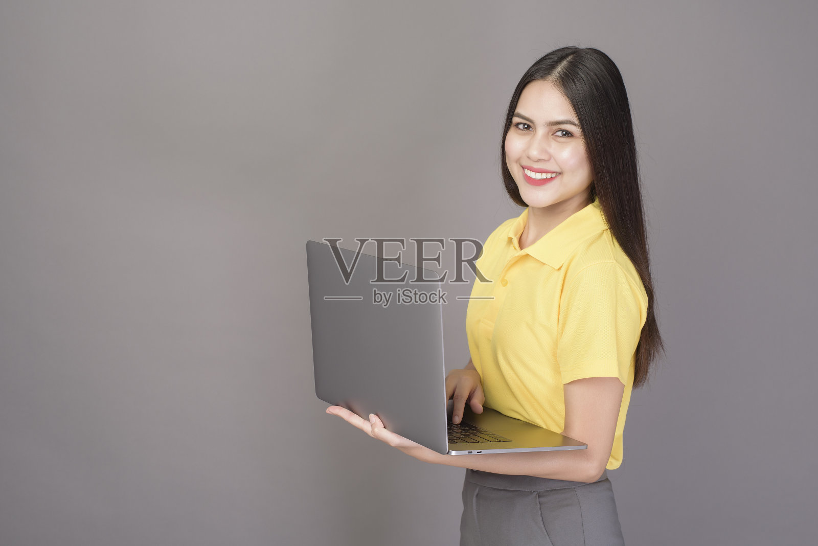 年轻自信的漂亮女人穿着黄色衬衫，拿着一个灰色背景的笔记本电脑照片摄影图片