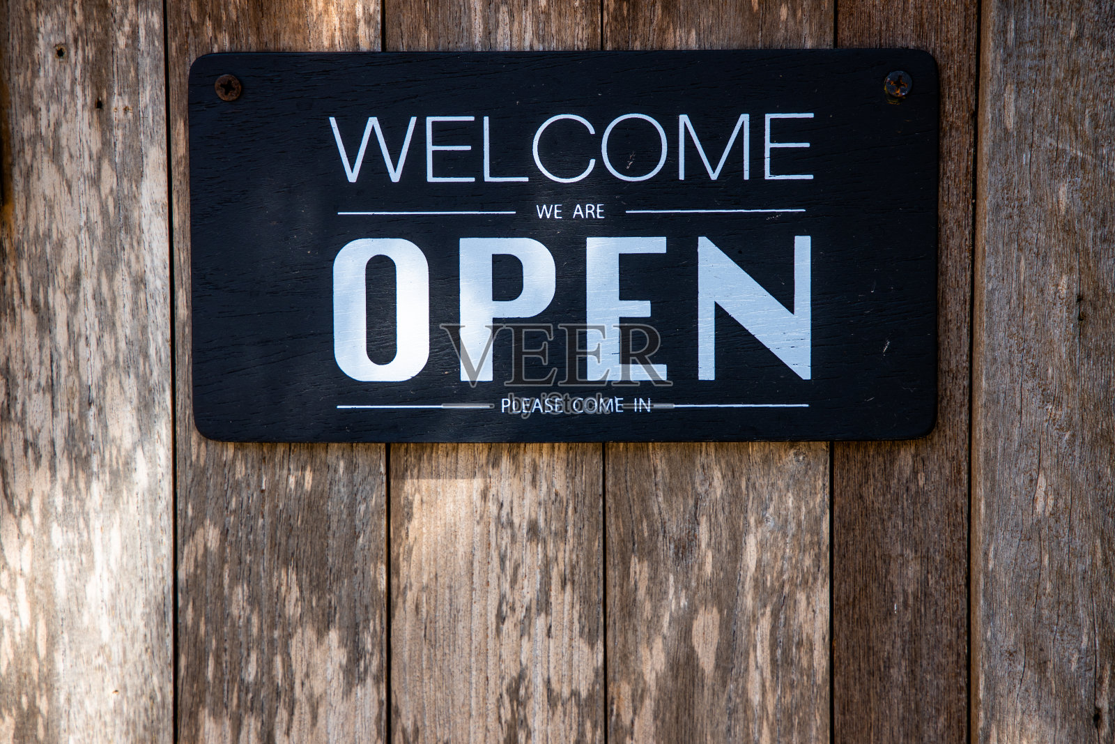 一个“欢迎，我们开门。”请用白色英文大写字母挂在旧木门上的黑色木制背景。照片摄影图片