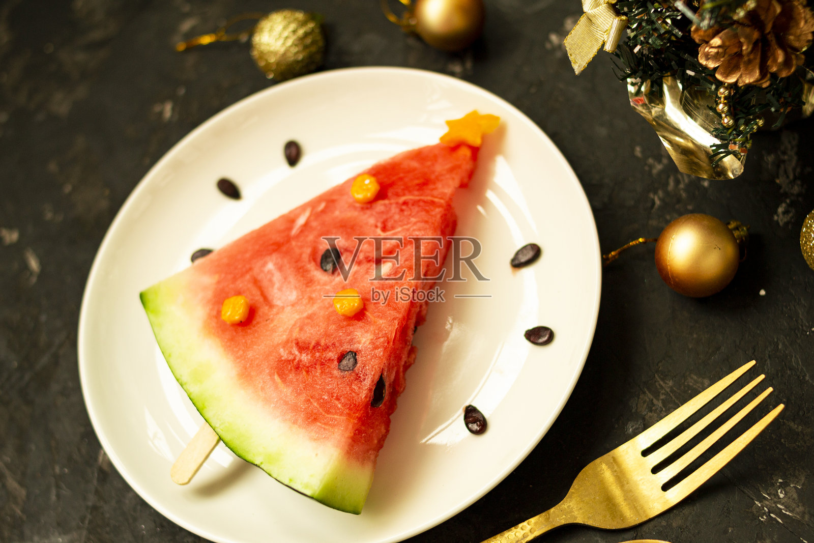 一块西瓜被装饰成白色盘子上的圣诞树。黑色混凝土背景，圣诞球和叉子勺子和装饰。给孩子们的早餐点子。新年，圣诞美食配西瓜，顶观。庆祝活动照片摄影图片