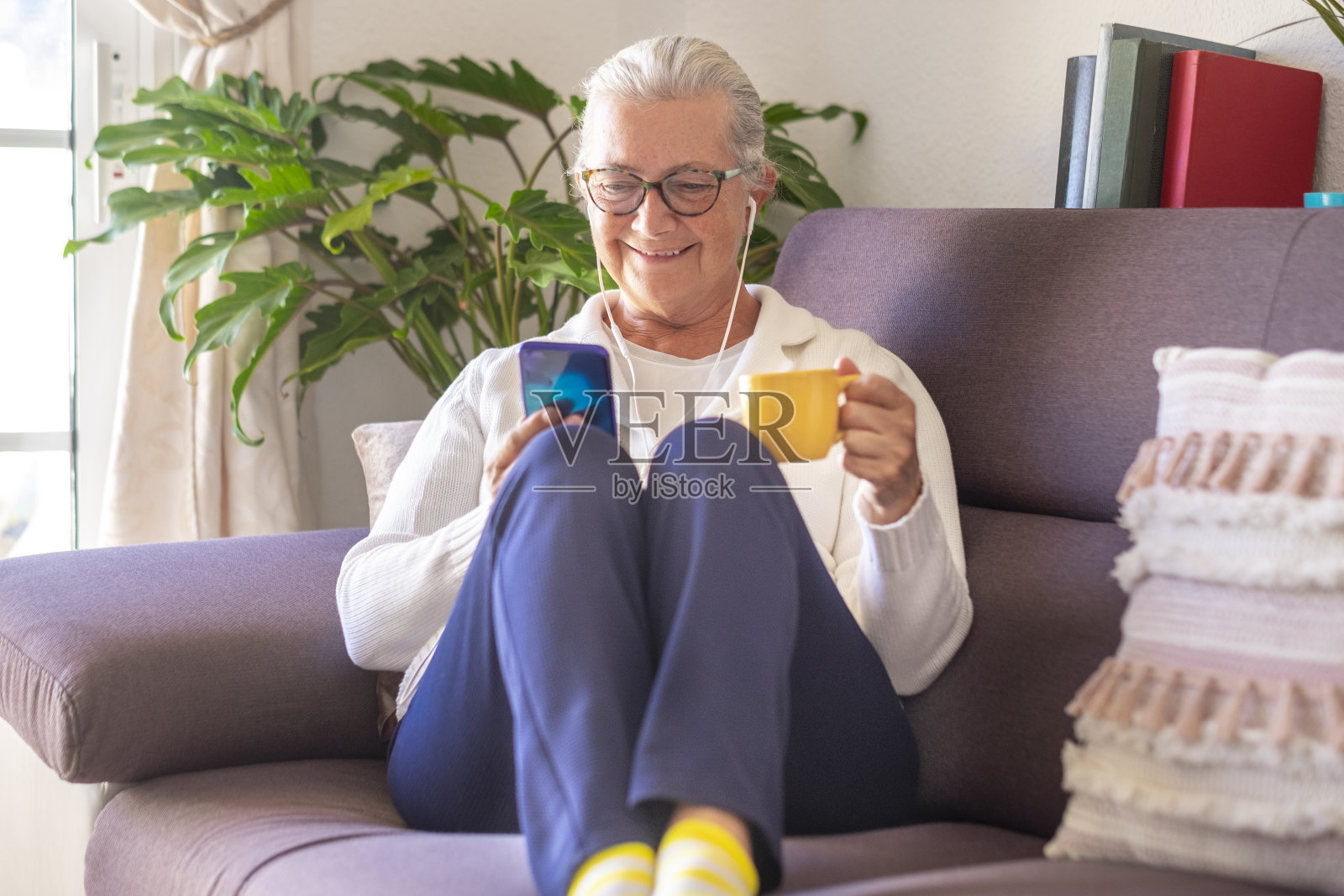微笑的老妇人坐在家里的沙发上用她的智能手机和朋友或家人聊天-女人白发，戴着眼镜耳机-活跃的社交和技术的人照片摄影图片