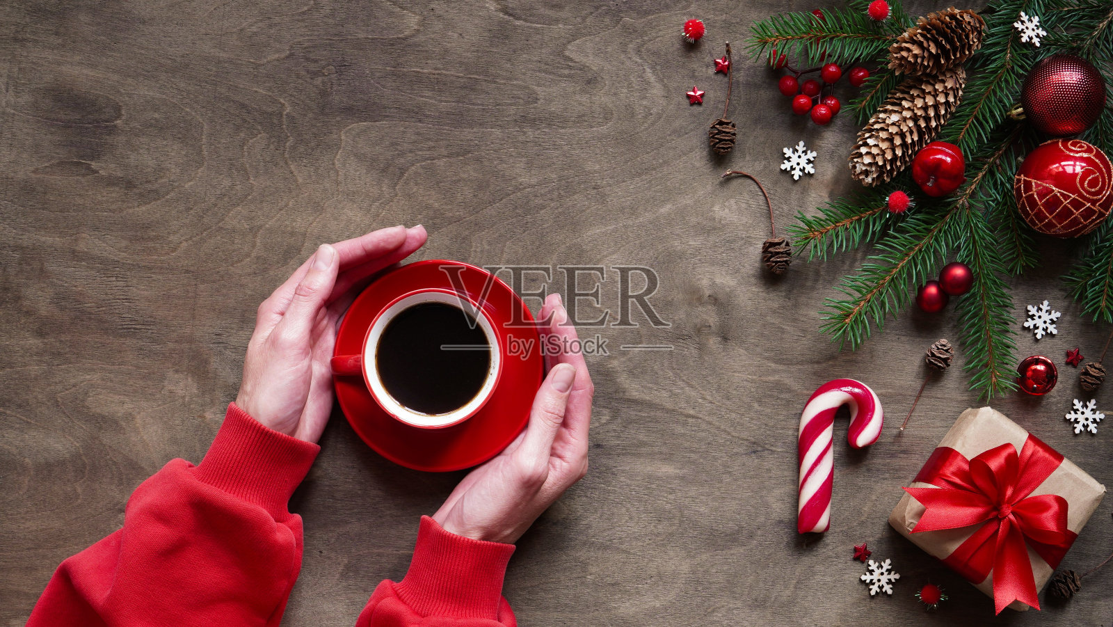 女人拿着红色的咖啡在圣诞装饰，球，棒棒糖，礼品盒，圣诞树的树枝，锥体和雪花在木制背景，平铺，复制空间。圣诞、新年贺卡概念照片摄影图片